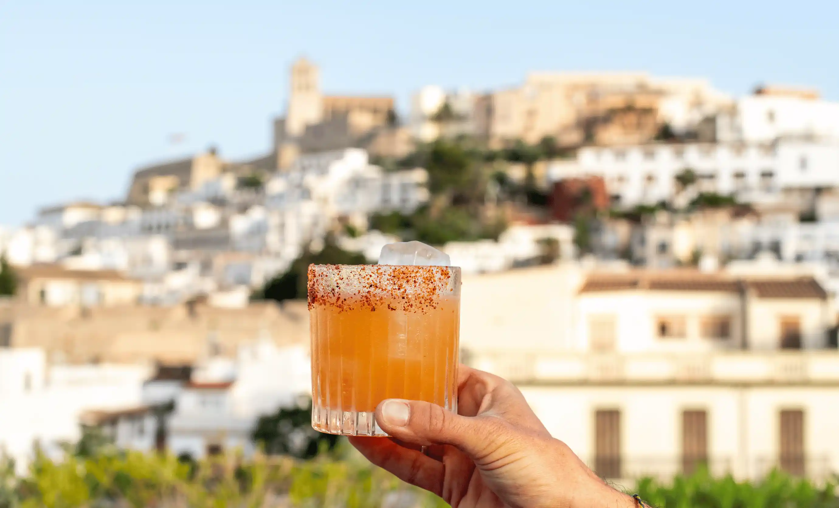 Hand holding an orange cocktail with a chili salted rim and ice cube, with a blurred town and castle in the background.