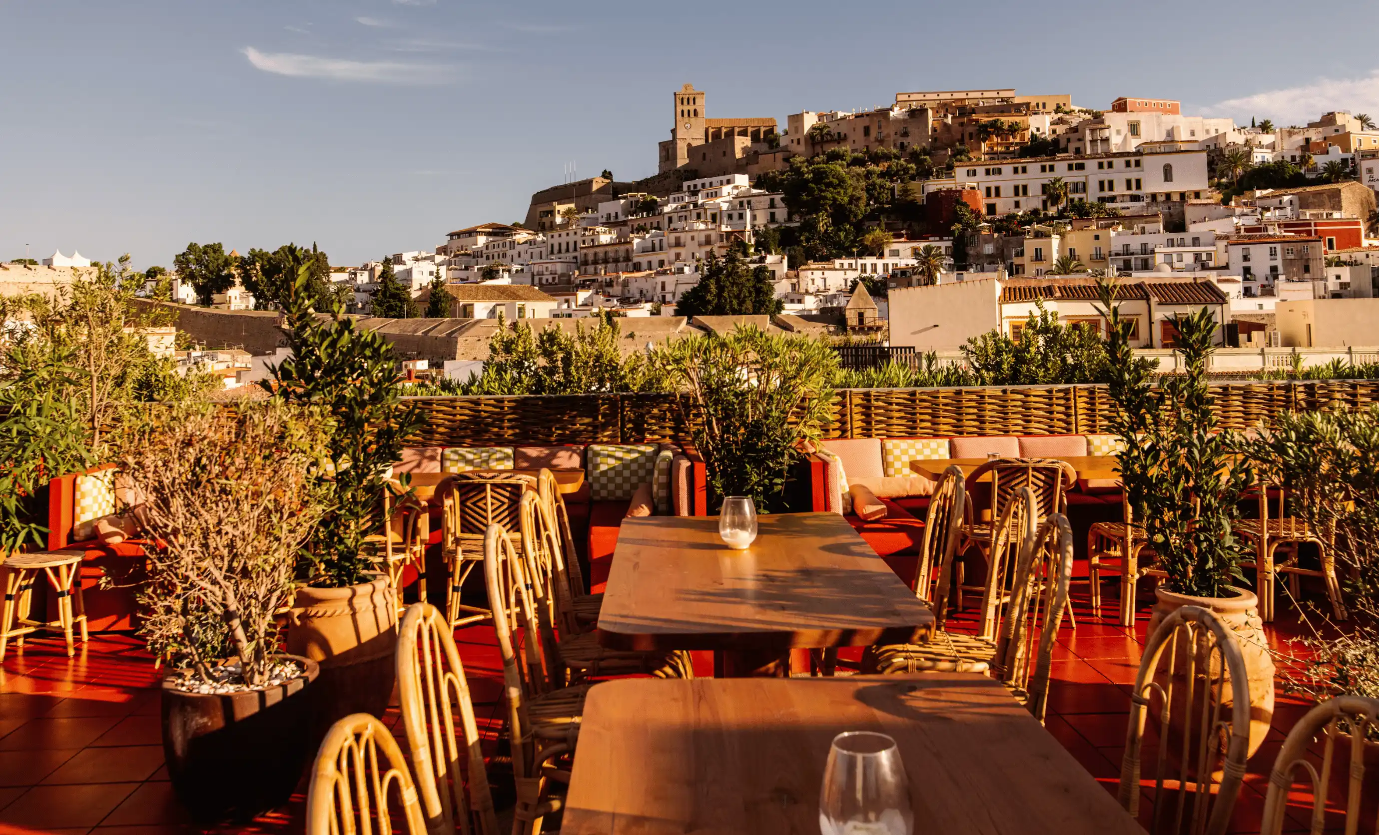 Sunlit outdoor terrace with wooden tables and wicker chairs overlooking a hillside town with white buildings and a historic clock tower.