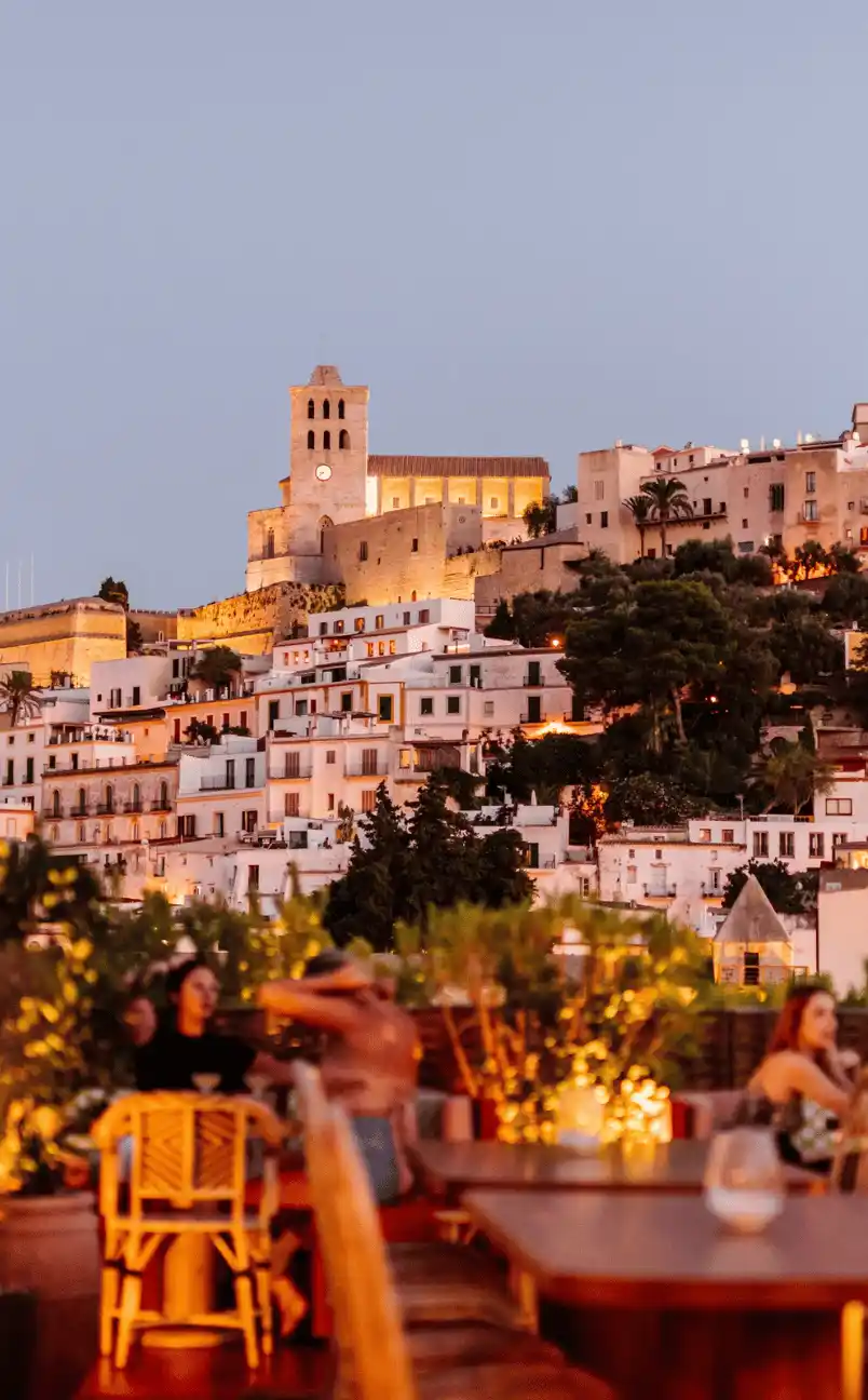 Evening view of a historic hilltop castle and white buildings with warm rooftop lights and people dining in the foreground.