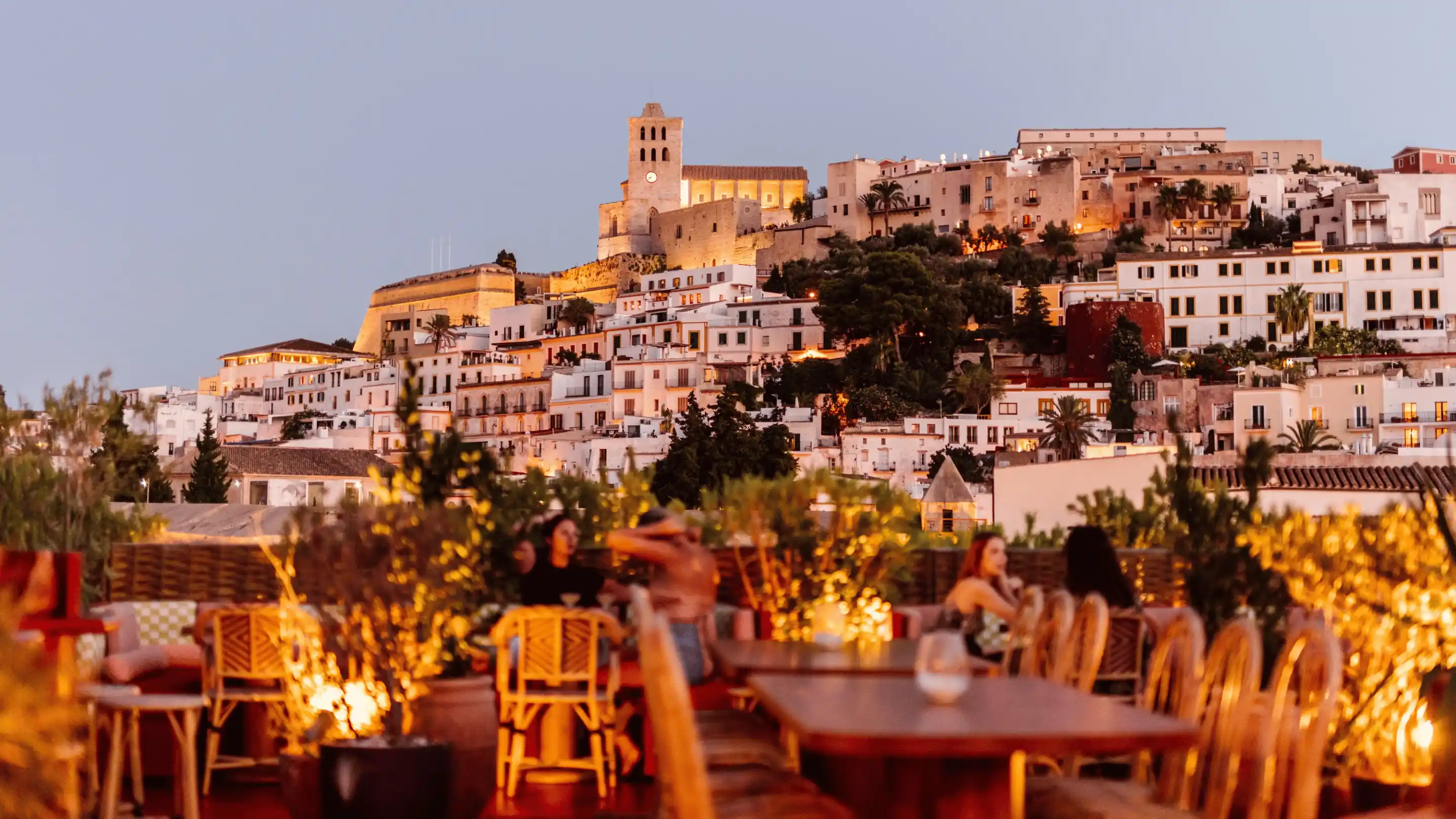 Evening view from a rooftop restaurant with wooden chairs and tables, overlooking a softly lit hillside town with historic buildings and a clock tower.