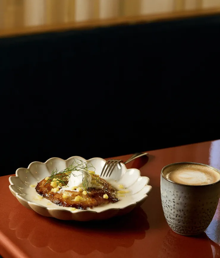 Dessert with cream and herbs on a white scalloped plate next to a ceramic cup of coffee on a red surface.