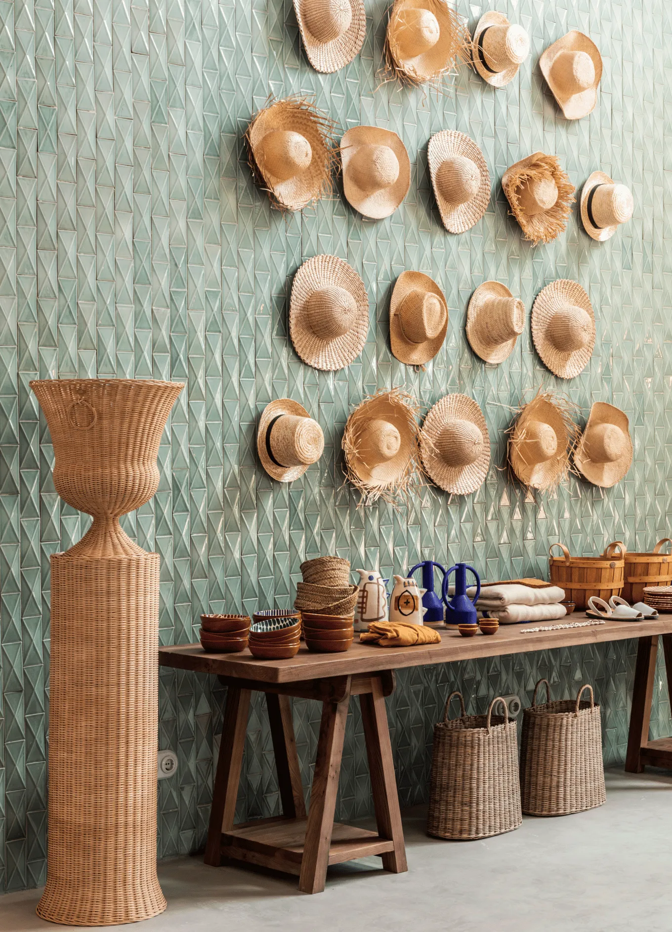 Wall decorated with multiple woven straw hats above a wooden table displaying woven baskets, ceramic jugs, bowls, and folded textiles.