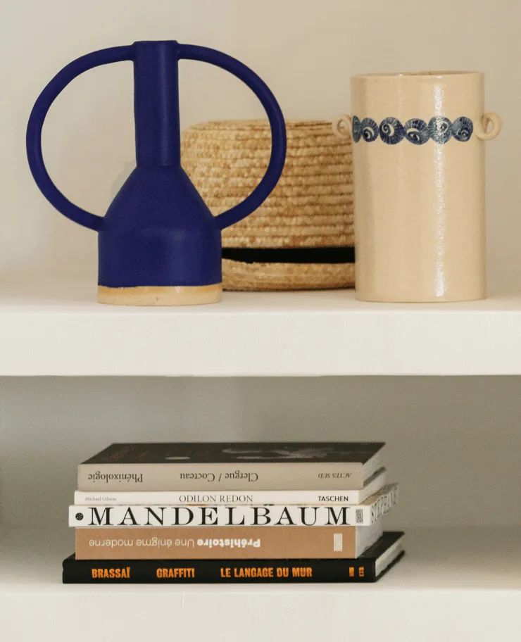 A beige ceramic vase with blue spiral patterns and a blue decorative object with circular handles on a shelf above a stack of art books.