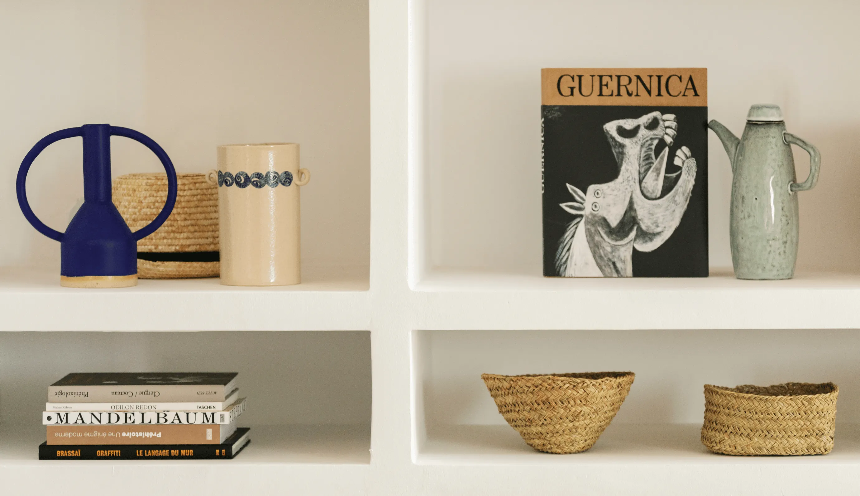 White bookshelf with decorative items including a blue ceramic vase, woven basket, beige vase, art book titled Guernica, green pitcher, stack of art books, and two woven baskets.