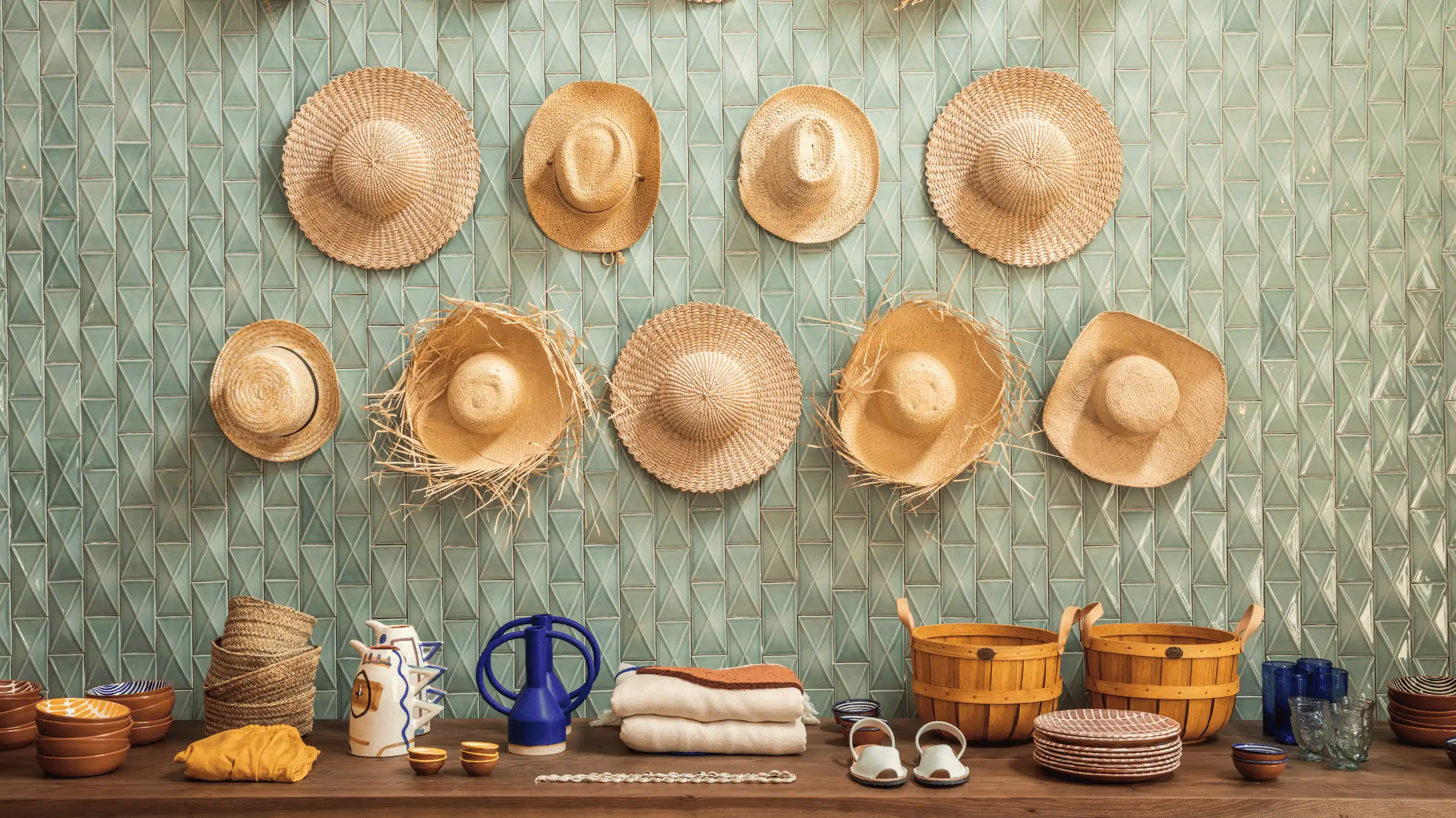 Display of various straw hats hanging on a green tiled wall above a wooden table with ceramic bowls, woven baskets, folded textiles, sandals, and glassware.