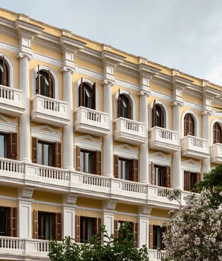 Yellow and white classic hotel facade with multiple arched windows and small balconies, framed by white columns and greenery in front.