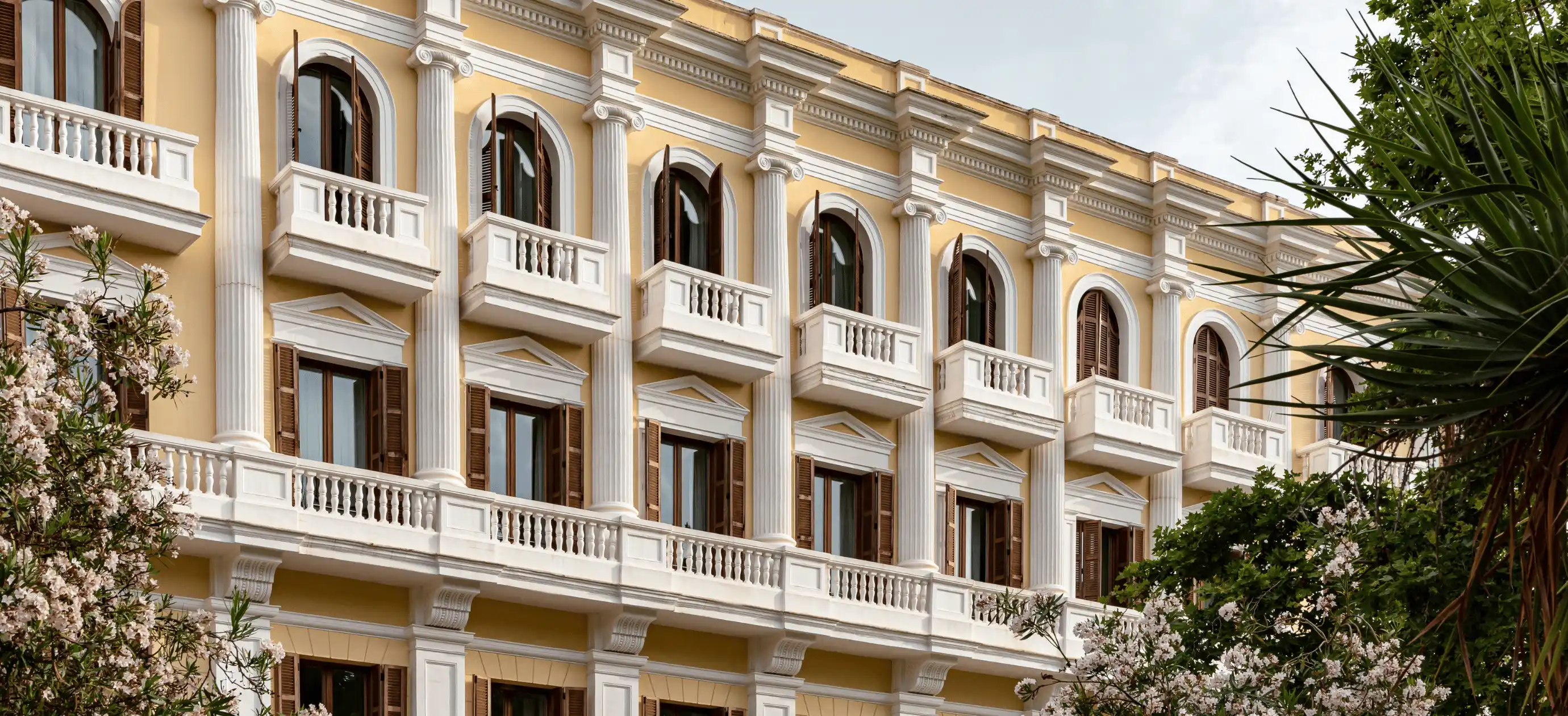 Yellow hotel building facade with white columns, arched windows with wooden shutters, and small balconies surrounded by greenery and flowering trees.