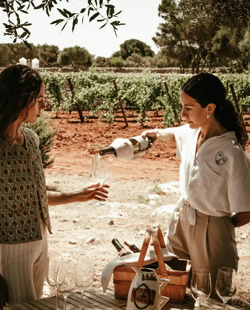 Two women by a table outdoors near a vineyard, one pouring white wine into a glass held by the other.