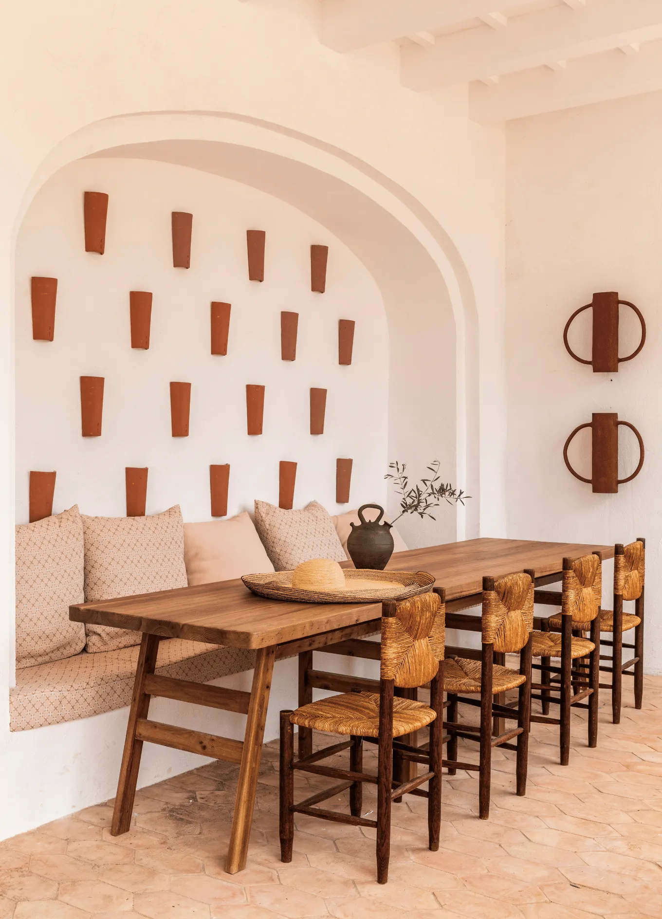 Cozy dining area with a long wooden table, woven chairs, a bench with patterned cushions, terracotta wall pots, and a straw hat on the table.