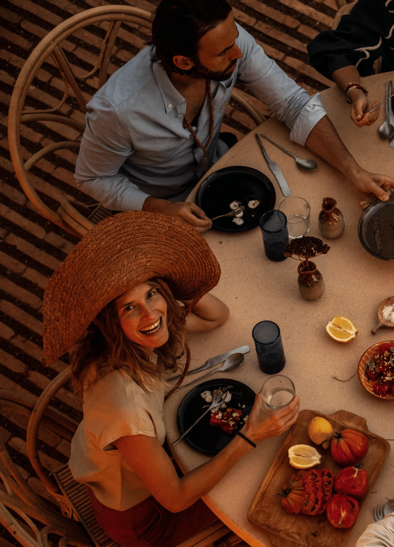Smiling woman in a large straw hat sitting at a table with food and drinks, joined by other people dining together.