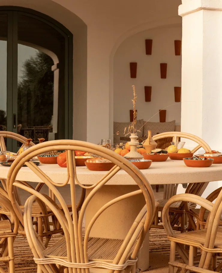 Outdoor dining area with a round beige stone table set with bowls of fruit and wicker chairs on a terrace with archways and a cushioned bench in the background.