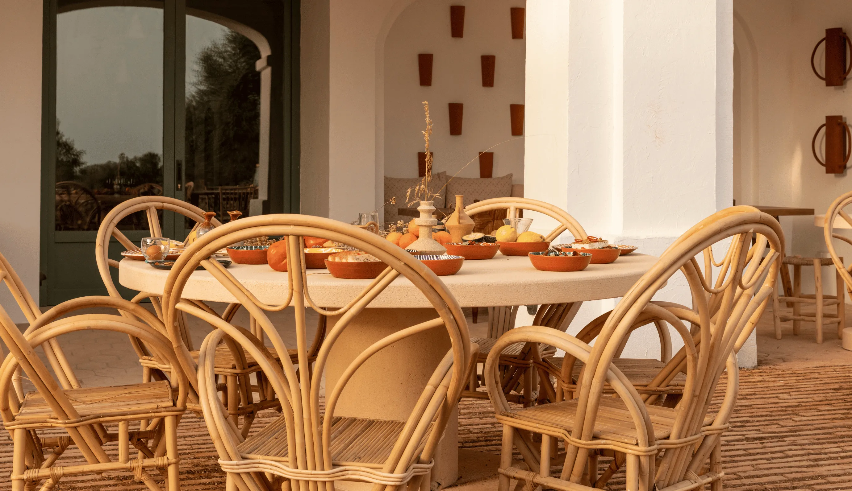 Outdoor round table set with wicker chairs on a terrace decorated with bowls, fruit, and pottery vases.