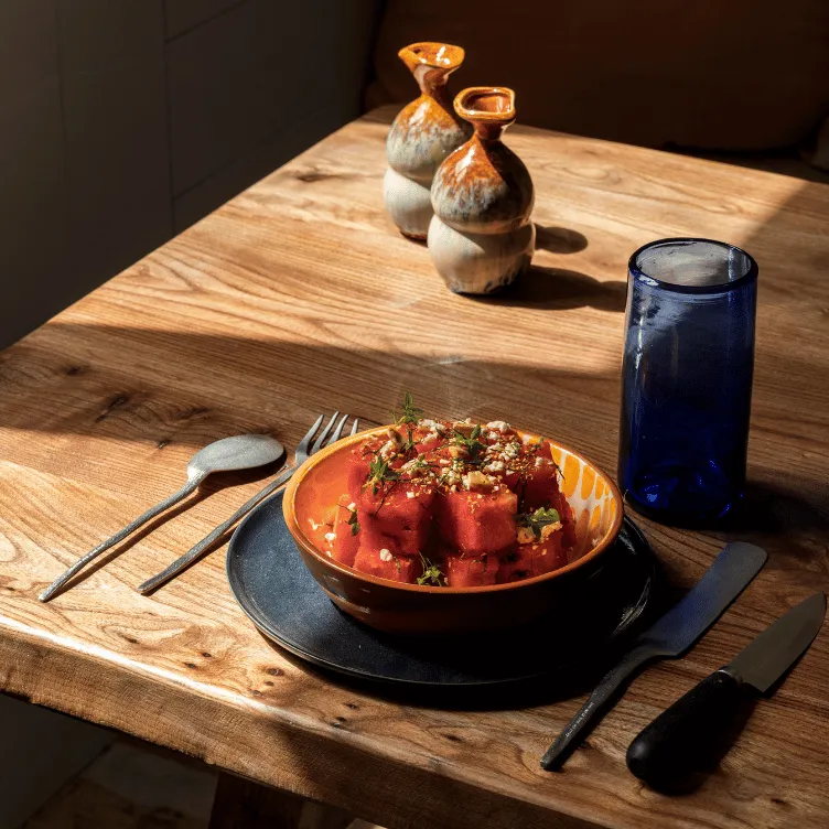 Bowl of cubed watermelon garnished with herbs and crumbs on a wooden table with a blue glass, two ceramic pitchers, and cutlery.