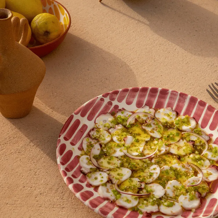 Plate of octopus carpaccio with sliced onions and green herb sauce on a red and white patterned plate, with a terracotta jug and bowl of lemons in the background.