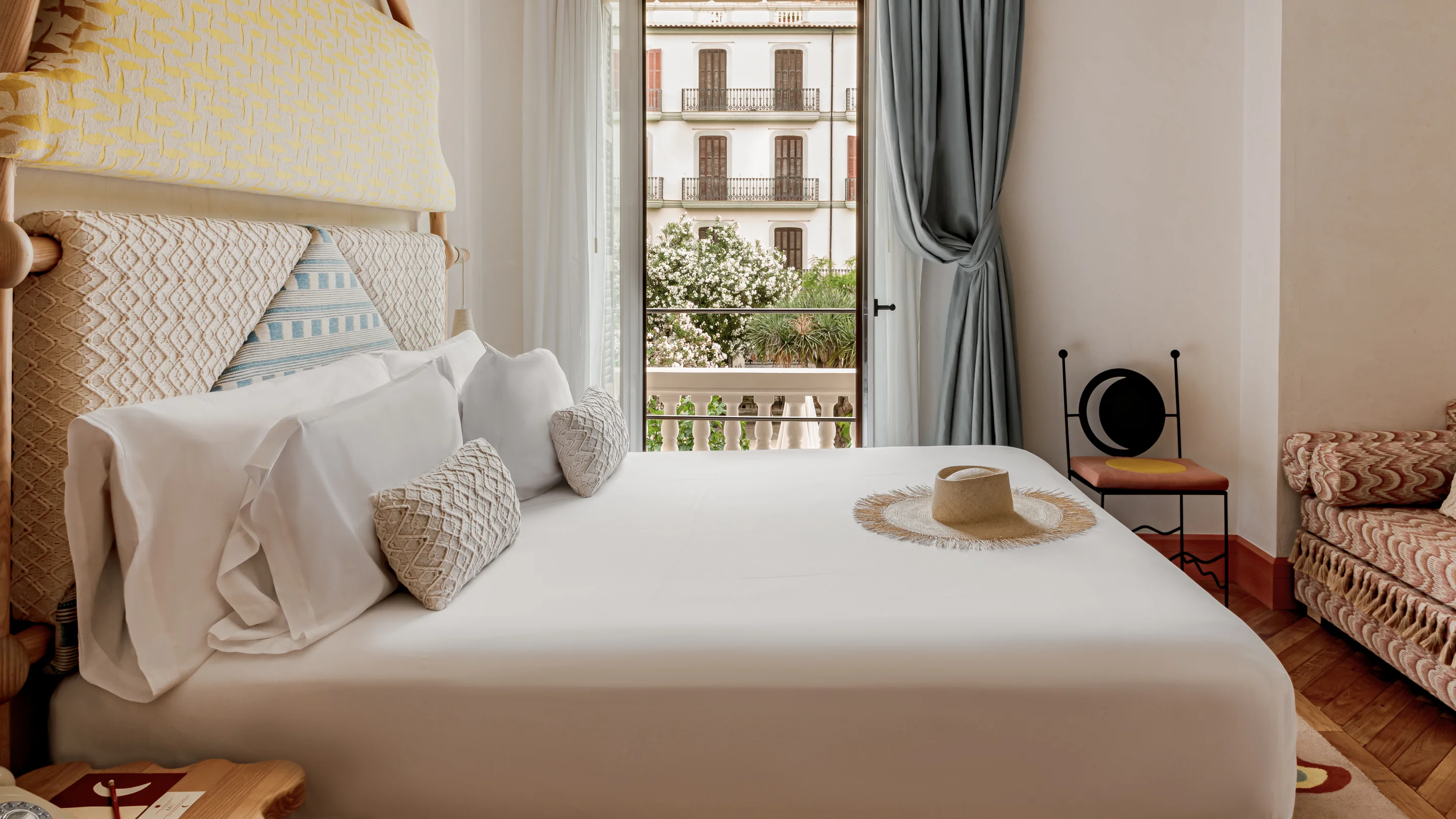 Neatly made bed with white linens and decorative pillows in a cozy room featuring a balcony door with a view of a building and greenery outside.