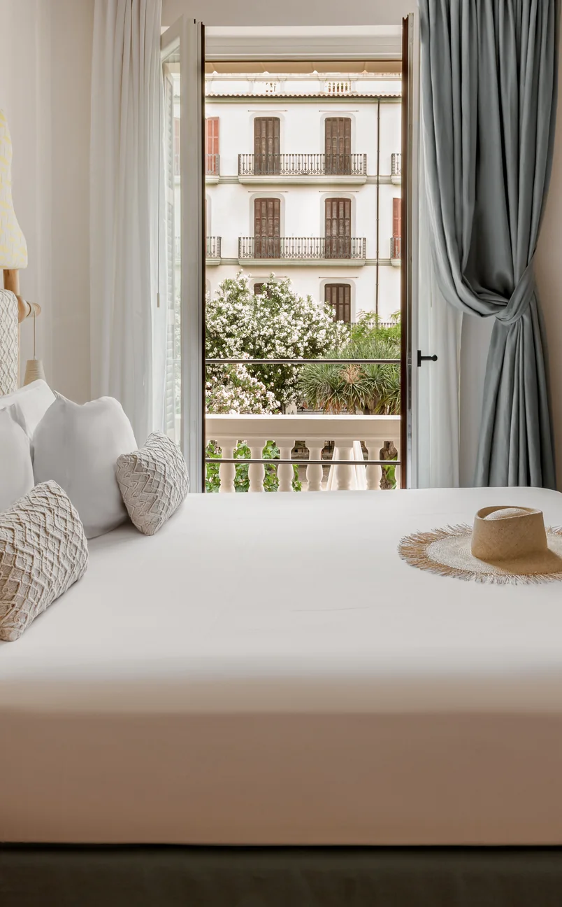 Modern hotel room with a neatly made bed, decorative pillows, a straw hat on the bed, and an open door leading to a balcony with flowering plants and a view of a classic building with balconies.