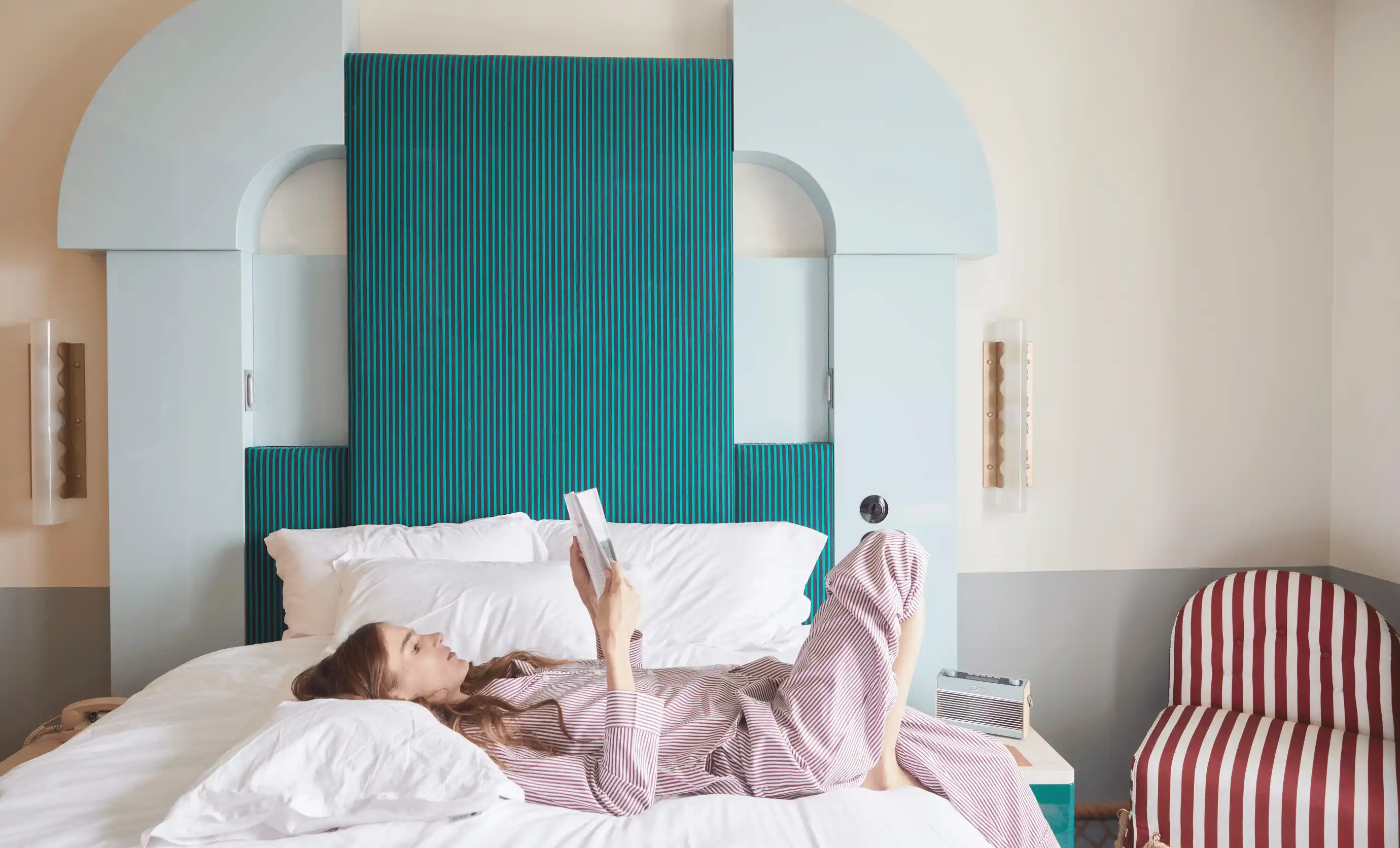 Woman in striped pajamas lying on a bed reading a book in a room with a teal and light blue headboard and a red-and-white striped chair.