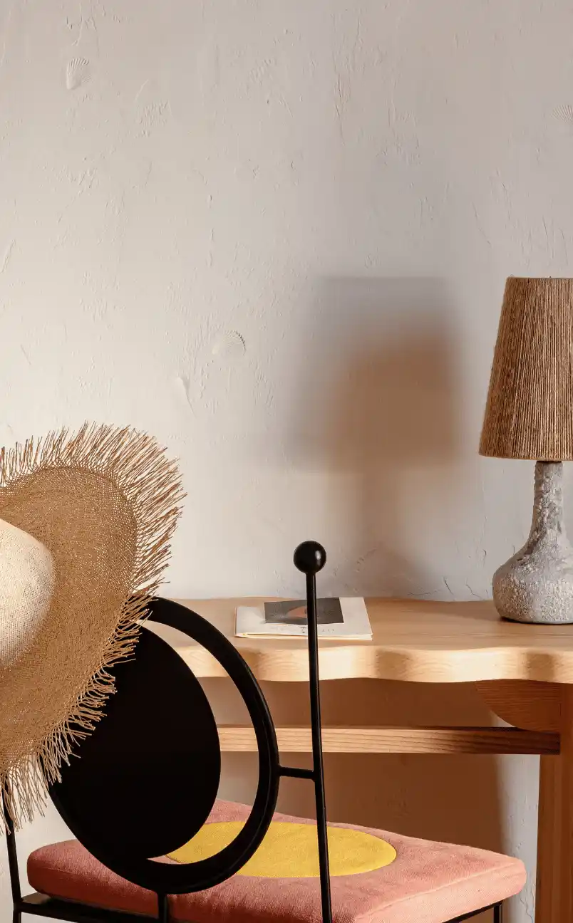 Sombrero de paja para el sol sobre una silla negra con un cojín naranja y amarillo, junto a una mesa de madera clara con una lámpara texturizada y un libro.
