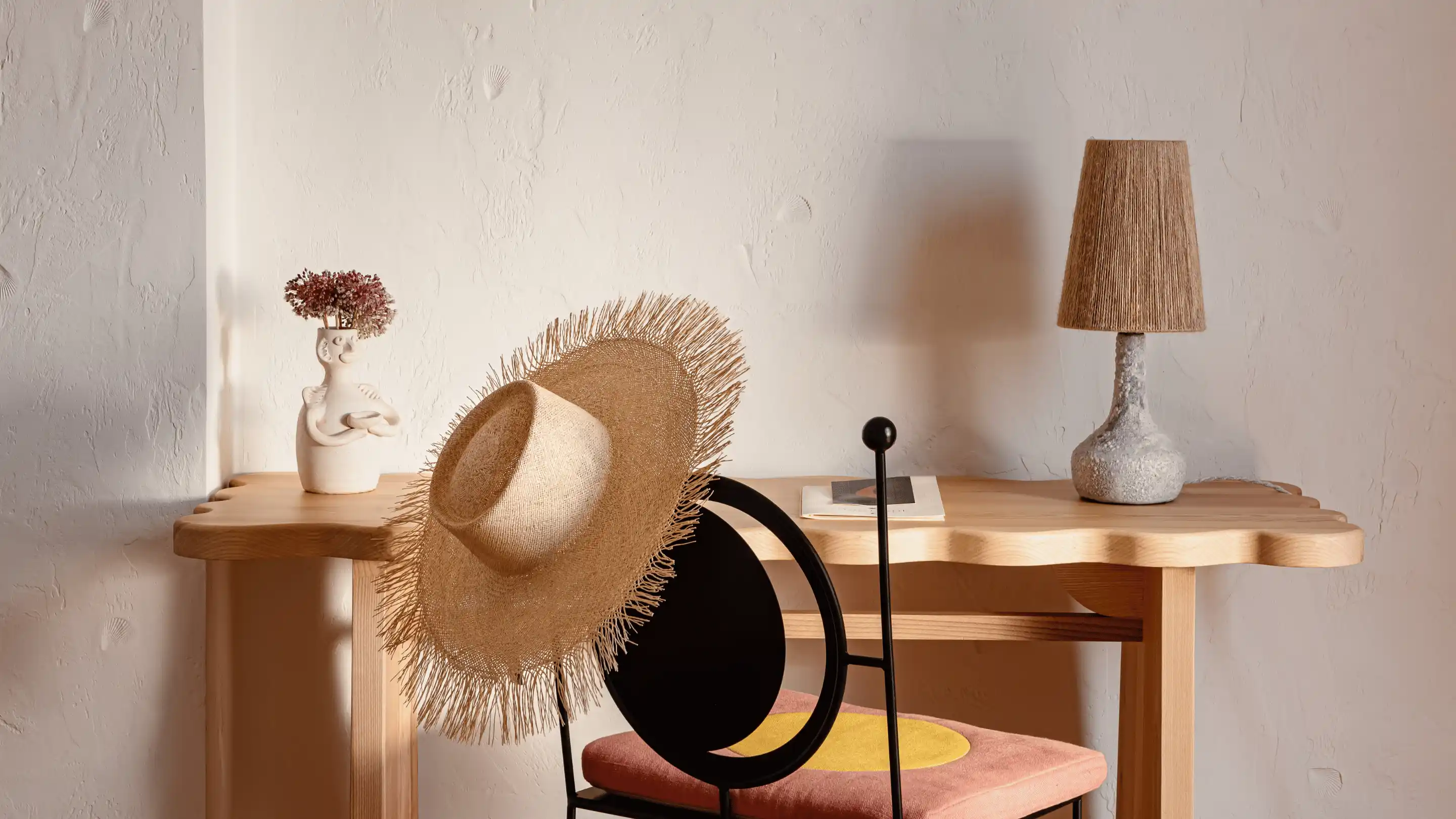 Sombrero de paja tejida que cuelga del respaldo de una silla de metal negro con cojín rosa y amarillo, junto a una mesa de madera con una lámpara texturizada y un jarrón de cerámica con flores secas.