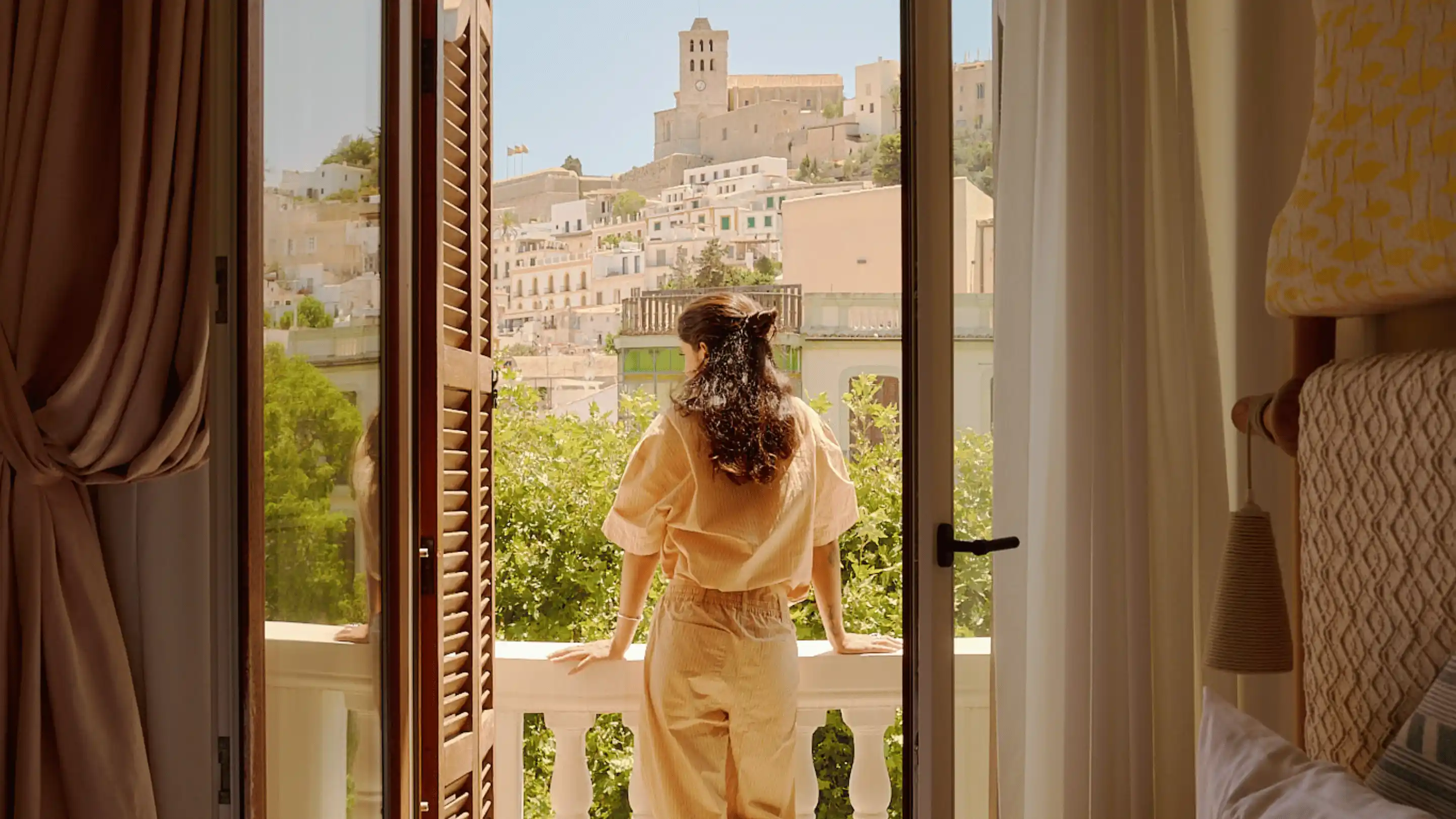 Femme en vêtements beiges debout sur un balcon regardant une ville à flanc de colline avec des bâtiments blancs et une tour d'horloge.