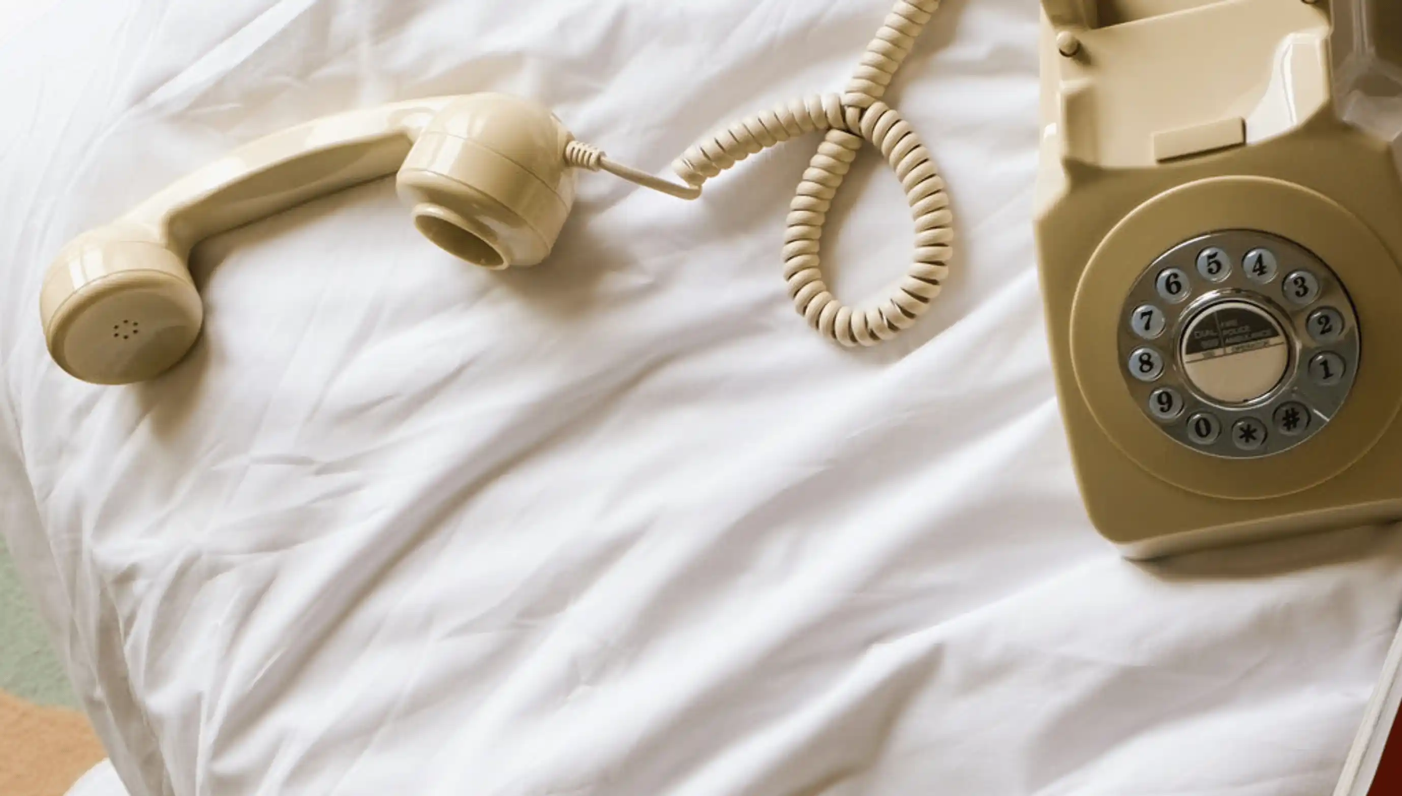Beige rotary dial telephone with handset off the hook resting on a white bed sheet.