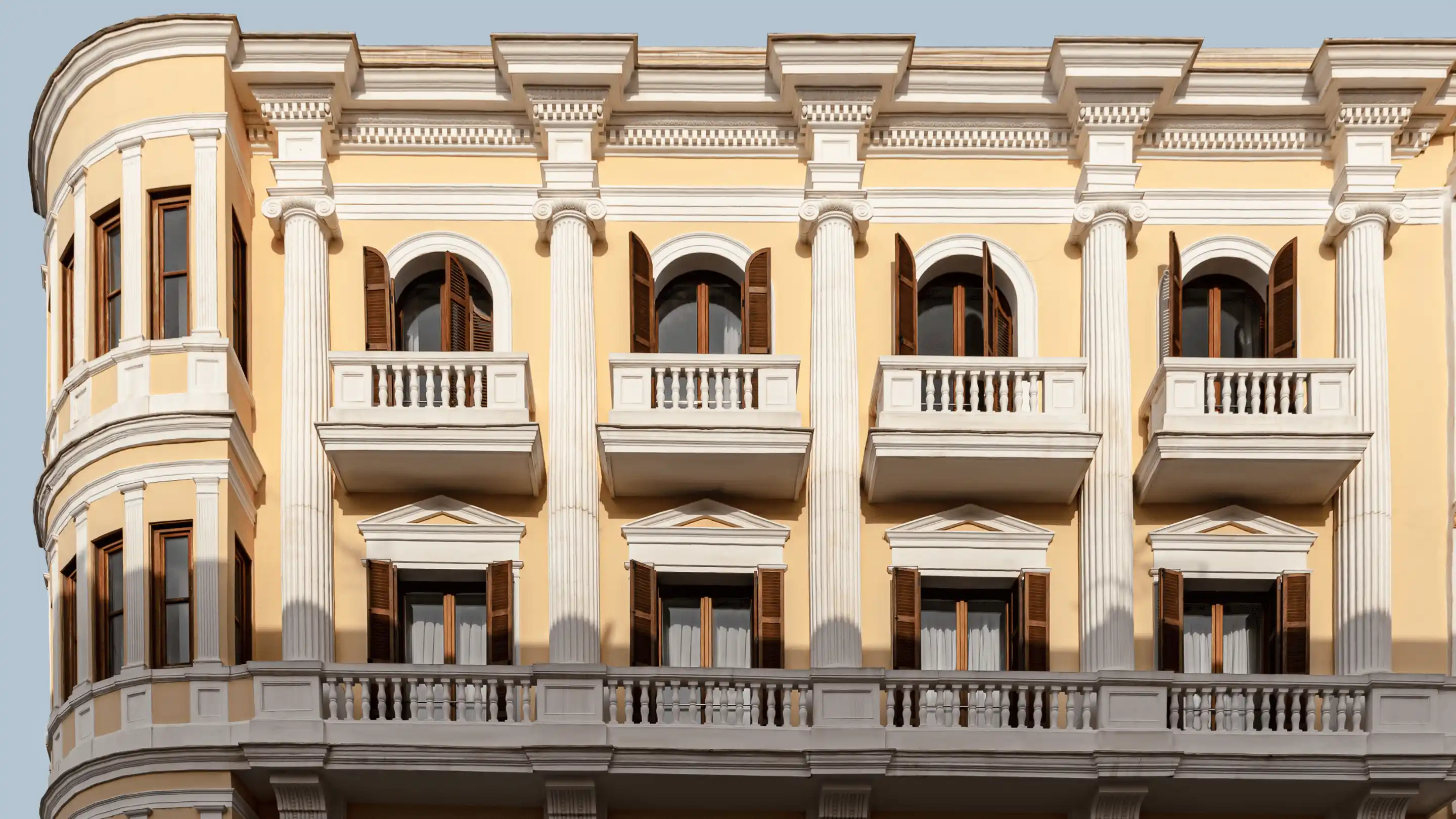 Front facade of a yellow and white classical building with balconies, columns, and wooden window shutters.