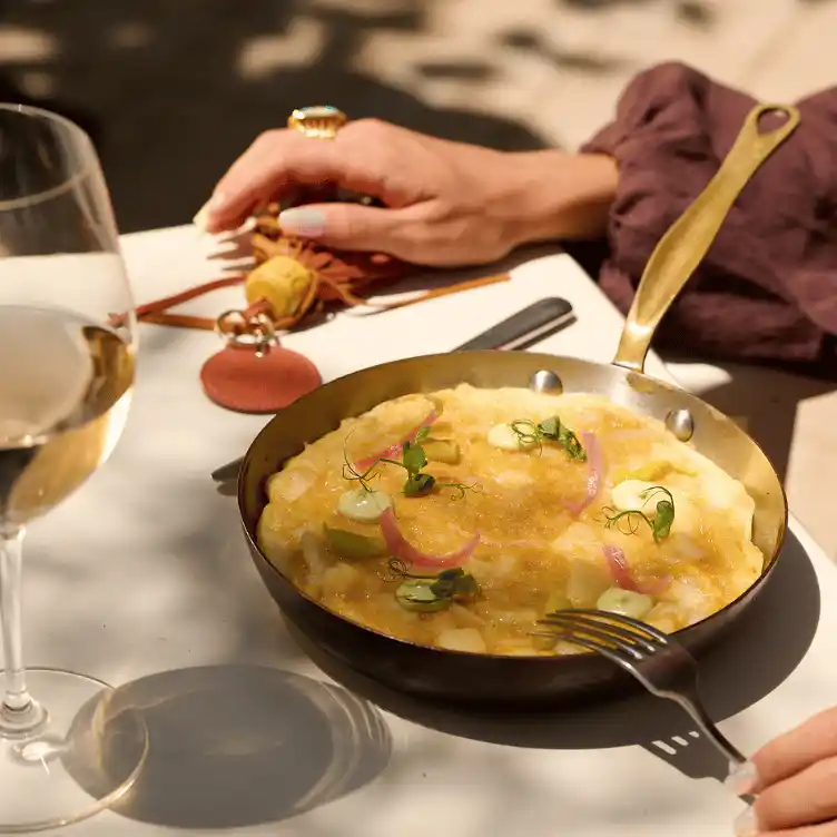 Skillet with gourmet dish garnished with microgreens and pickled onions on a white table, accompanied by a glass of white wine and a person holding a fork.
