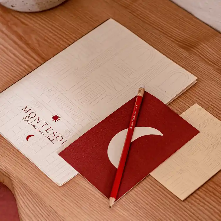 Wooden desk with a Montesol Experimental branded folder, a red notebook with a white crescent shape, and a red Montesol pencil on top.