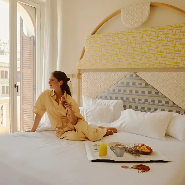 Woman in beige clothing sitting on a bed by a window, enjoying a croissant with breakfast tray holding juice, coffee, and fruit on the bed.