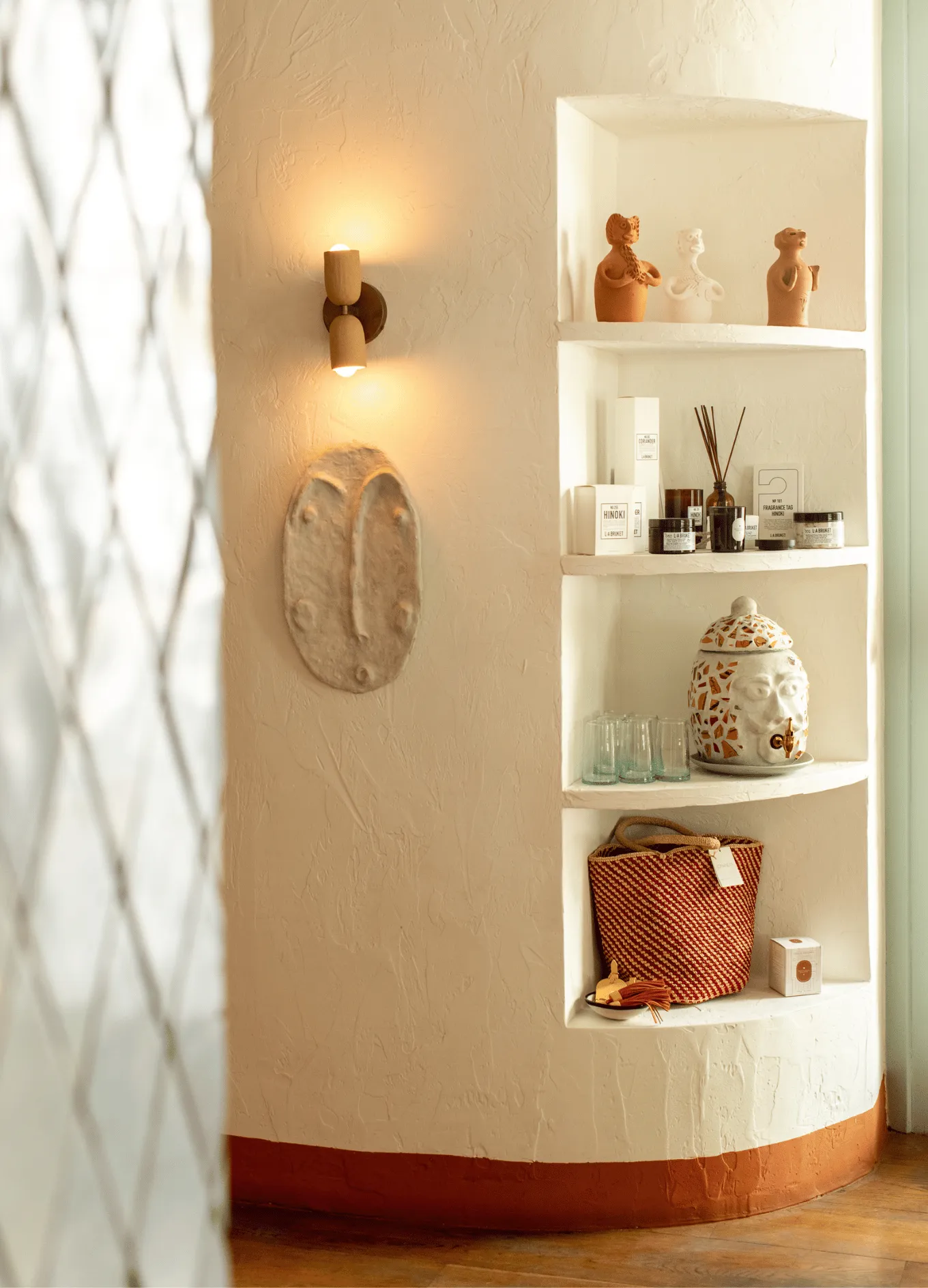 Curved wall niche with three shelves displaying clay figurines, candles, a decorative urn, drinking glasses, and a woven basket, beside a wall light and mask decoration.