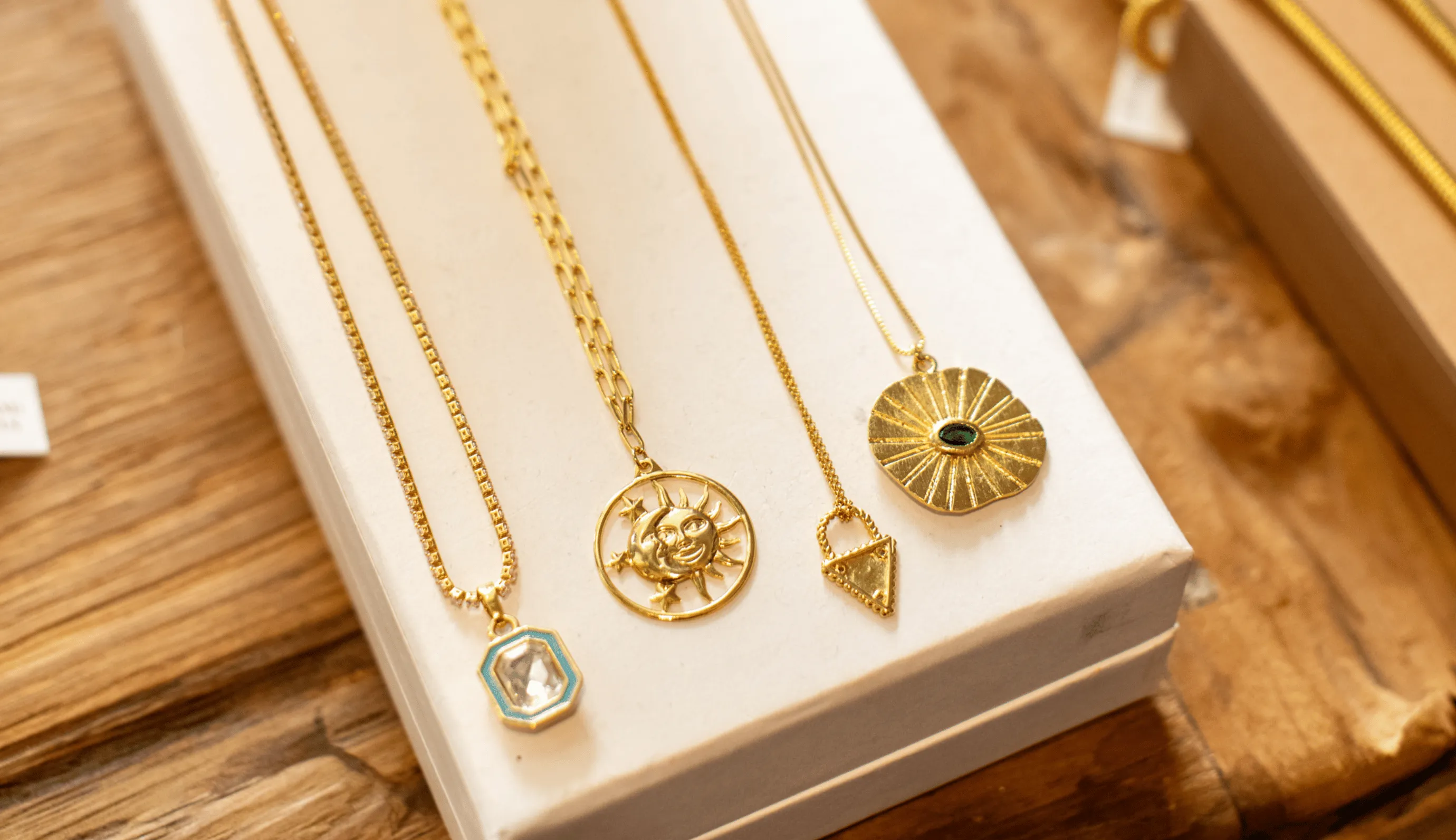 Four gold necklaces with unique pendants displayed on a white box on a wooden surface.