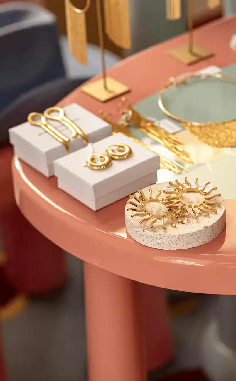 Gold-colored sun-shaped earrings displayed on a round terrazzo holder on a pink table alongside other gold earrings and jewelry pieces.