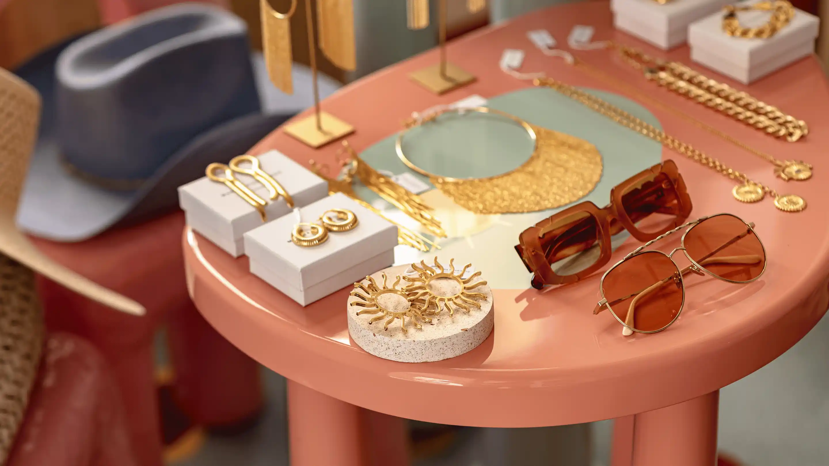 Display of gold jewelry including earrings, bracelets, and necklaces, alongside two pairs of brown tinted sunglasses on a round pink table with a gray hat in the background.