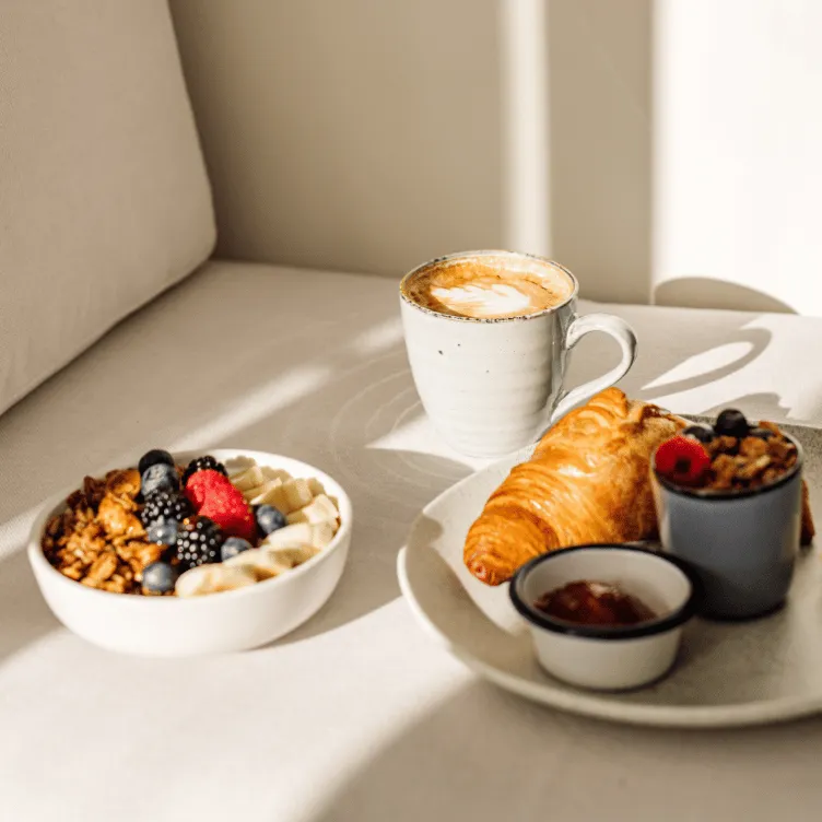 Breakfast setup with a cup of latte, a croissant, a small bowl of jam, a cup of granola with berries, and a bowl of sliced bananas and mixed berries on a white surface.