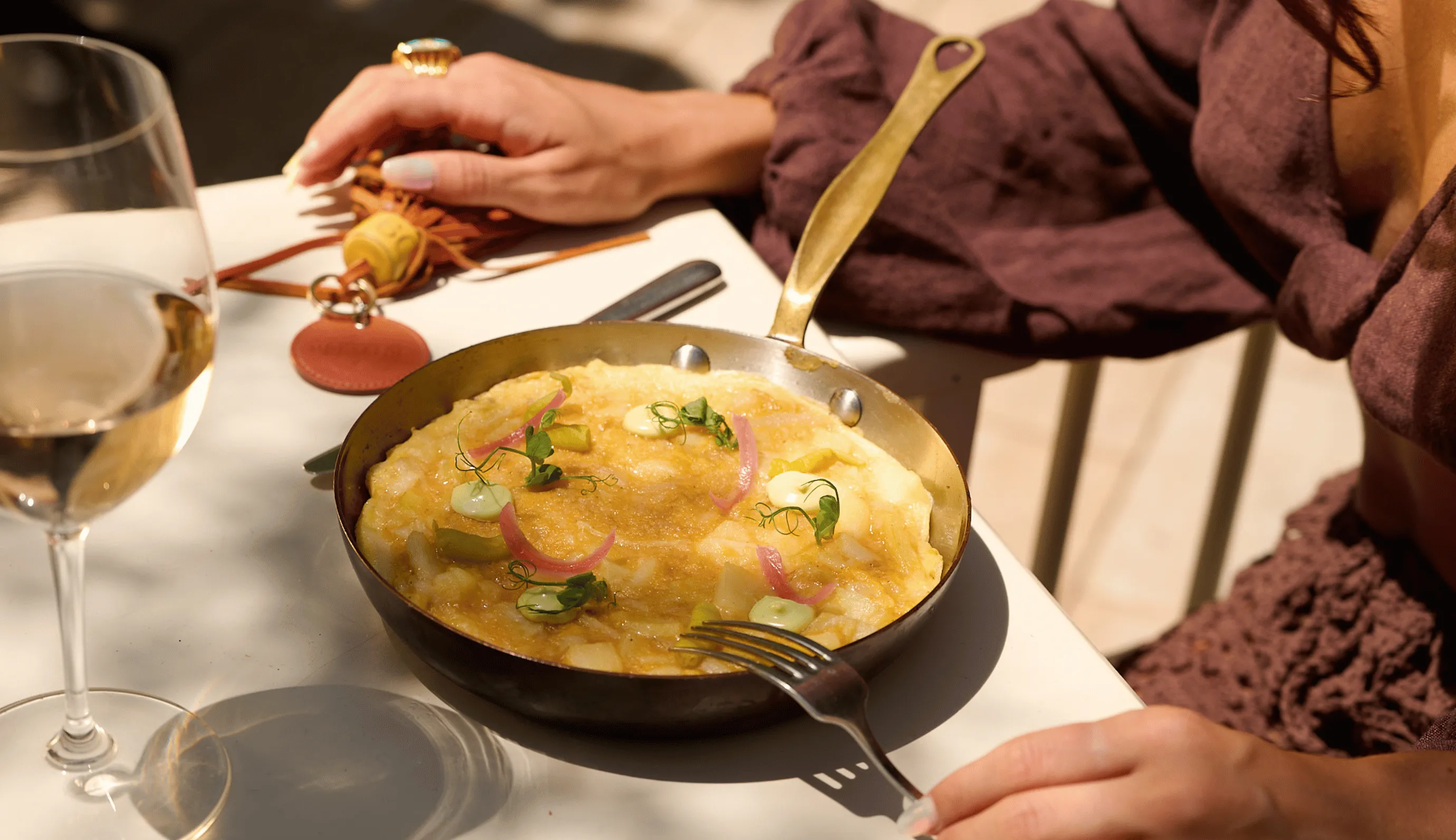Person in brown outfit holding a fork next to a frying pan with an omelet garnished with herbs and pickled onions on a white table with a glass of white wine.