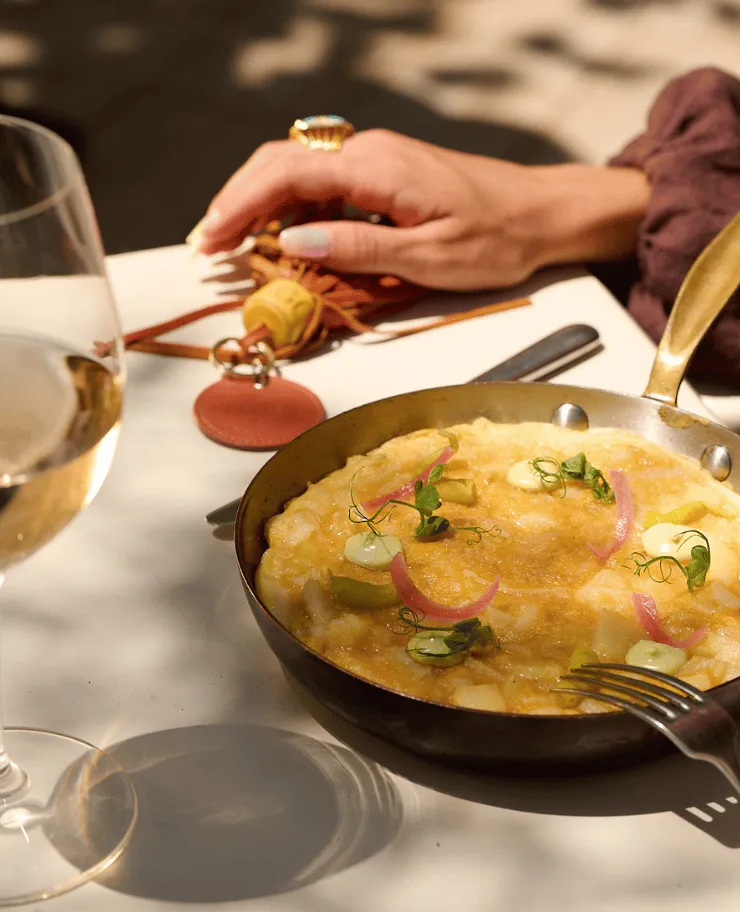 Pan with a creamy yellow dish garnished with herbs and pickled onions on a white table next to a glass of white wine and a hand resting on a leather handbag.