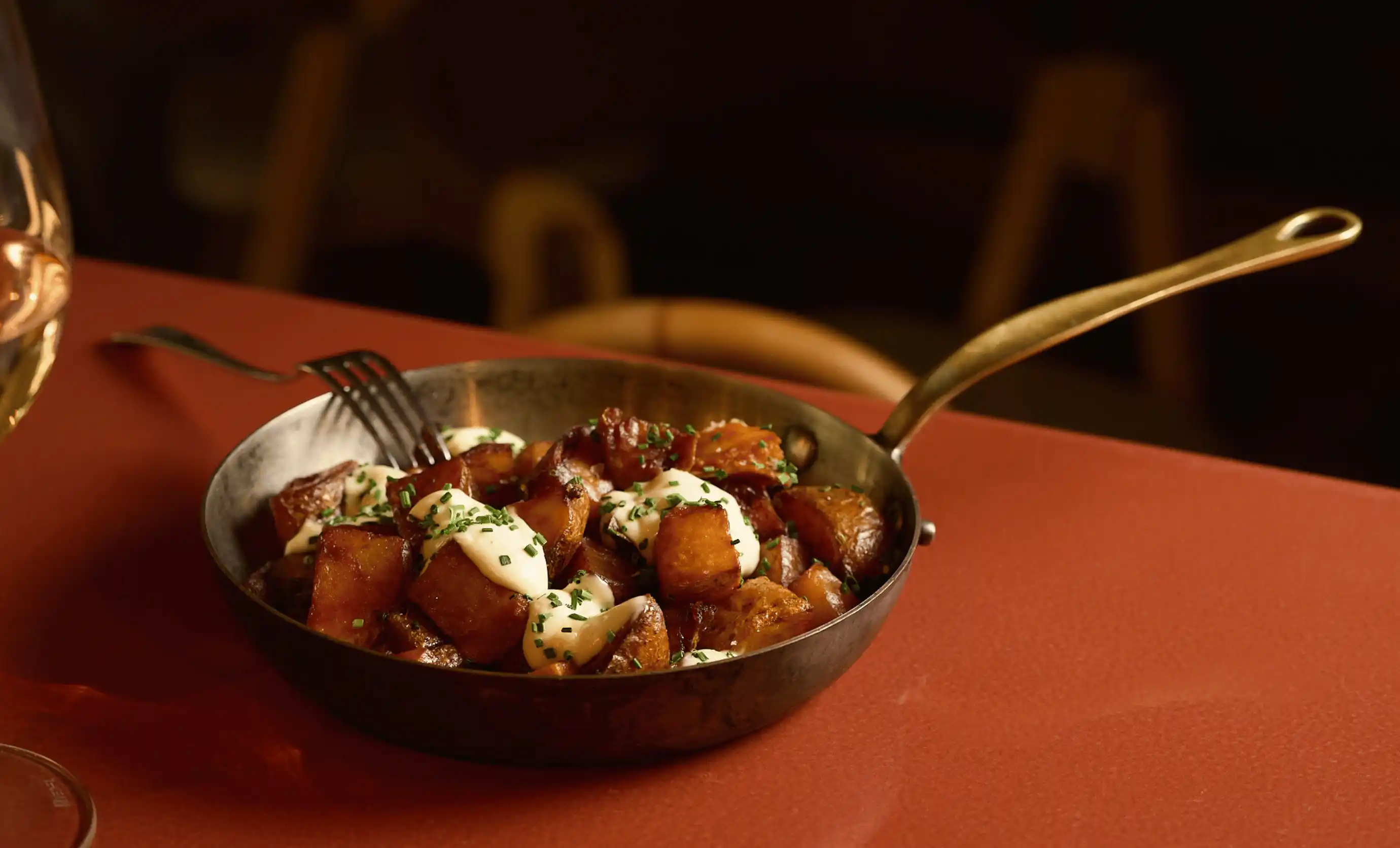 Small skillet with roasted potatoes topped with sour cream and chopped chives on a reddish table.