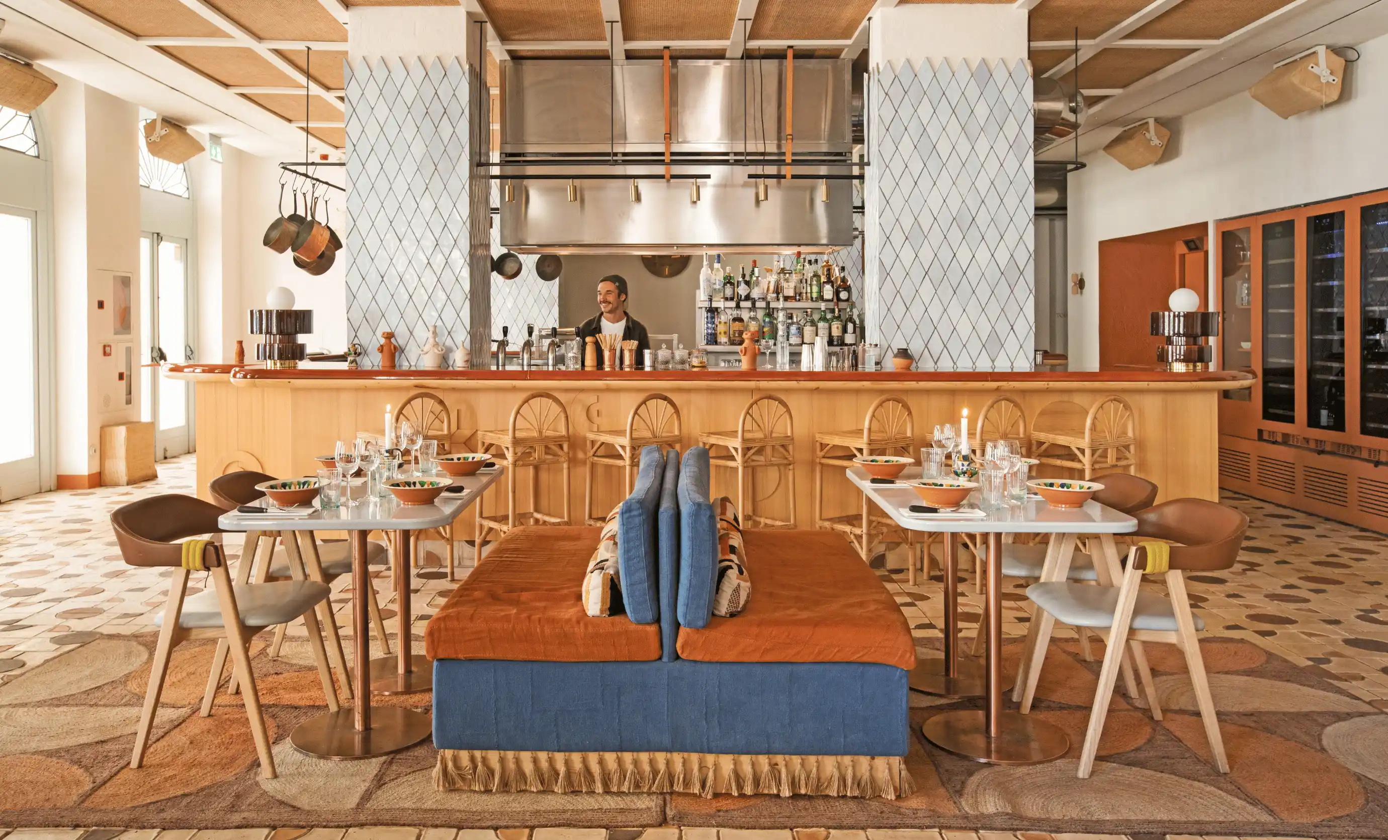 Modern restaurant interior with a central double-sided blue and orange cushioned bench, white marble tables set with bowls and glasses, wooden chairs, and a bar with a smiling bartender in the background.