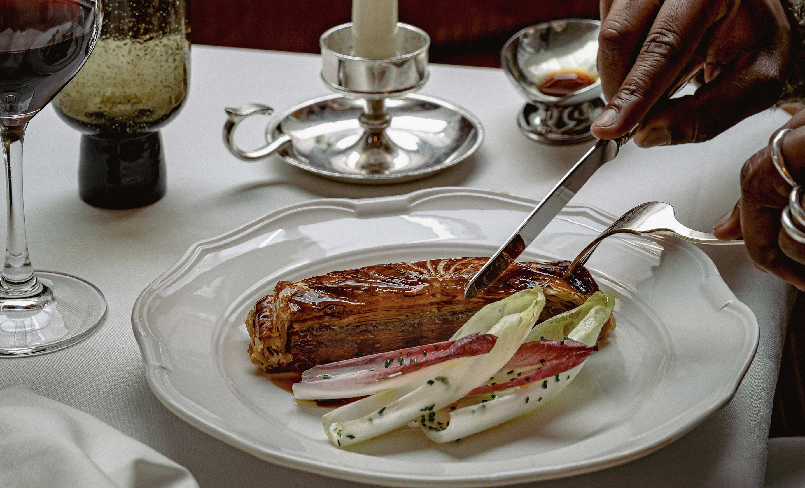 Hands cutting a golden puff pastry on a white plate garnished with endive leaves, next to a glass of red wine on a white tablecloth.