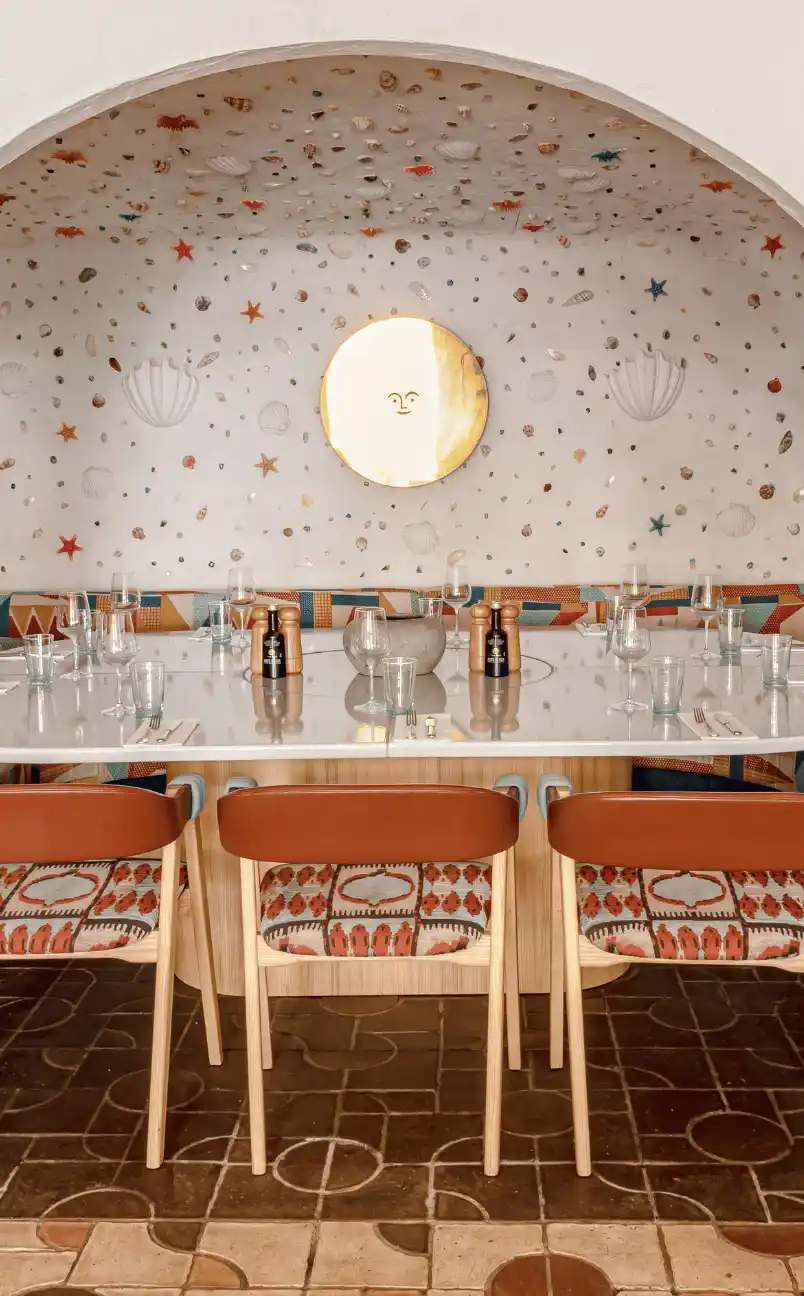 Dining table set with glassware, utensils, and pepper mills beneath an arched wall decorated with seashells, starfish, and a sun face mirror.