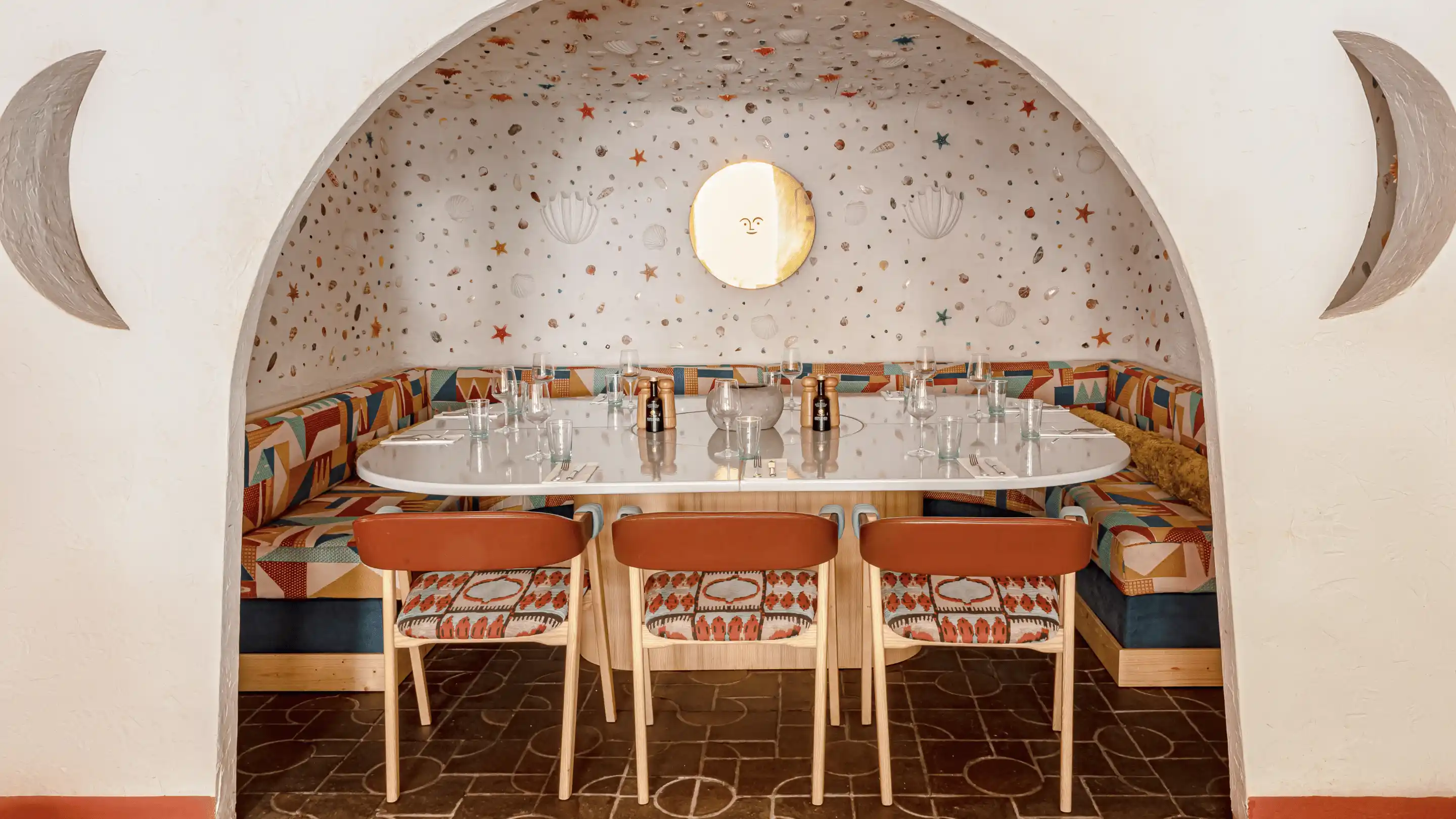 Curved white dining booth with colorful patterned cushions and three wooden chairs with leather backs under an arched alcove with moon shapes and a round mirror on a wall decorated with seashells and starfish.