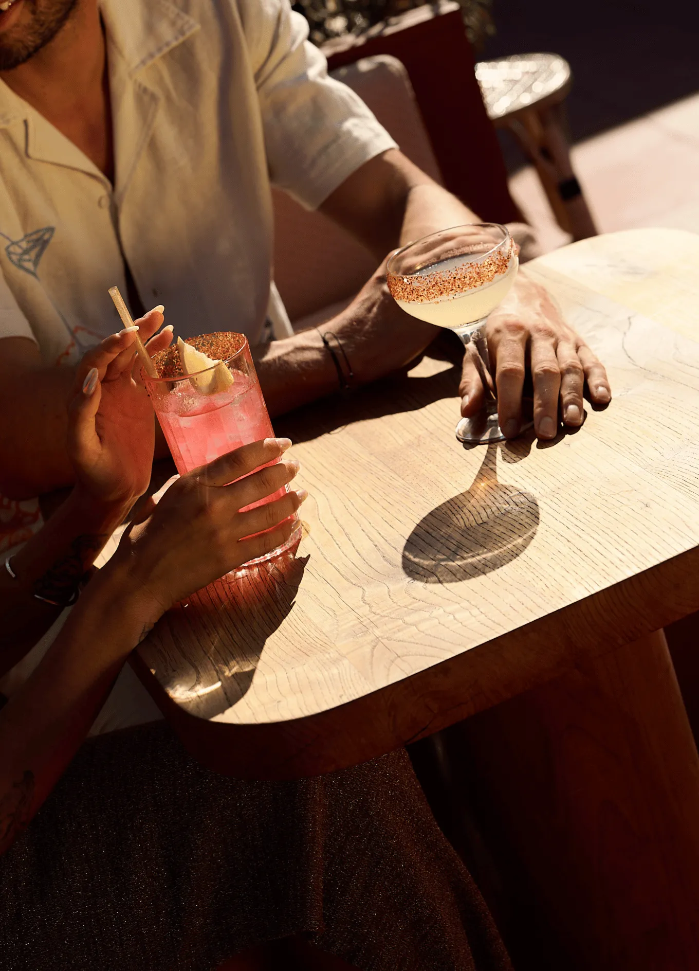 Two people at a wooden table holding cocktails, one with a pink drink and lemon garnish, the other with a light-colored drink in a coupe glass with a chili-rimmed edge.