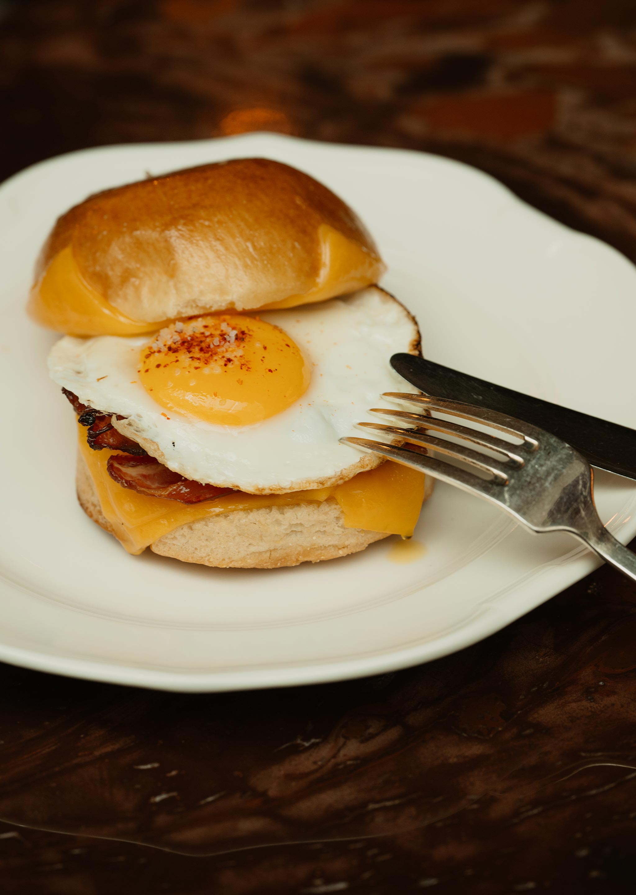 Breakfast sandwich with fried egg, cheddar cheese, and bacon on a bun, served on a white plate with fork and knife.