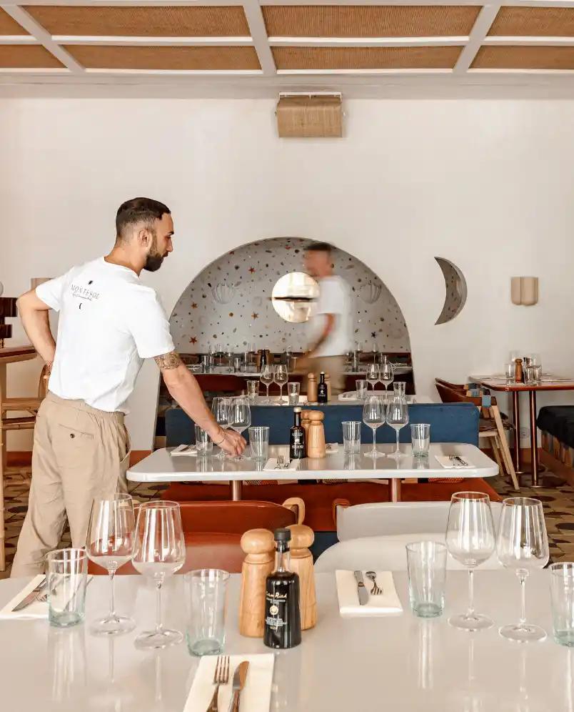 Man in beige pants and white shirt setting a modern restaurant table with glasses and condiments.