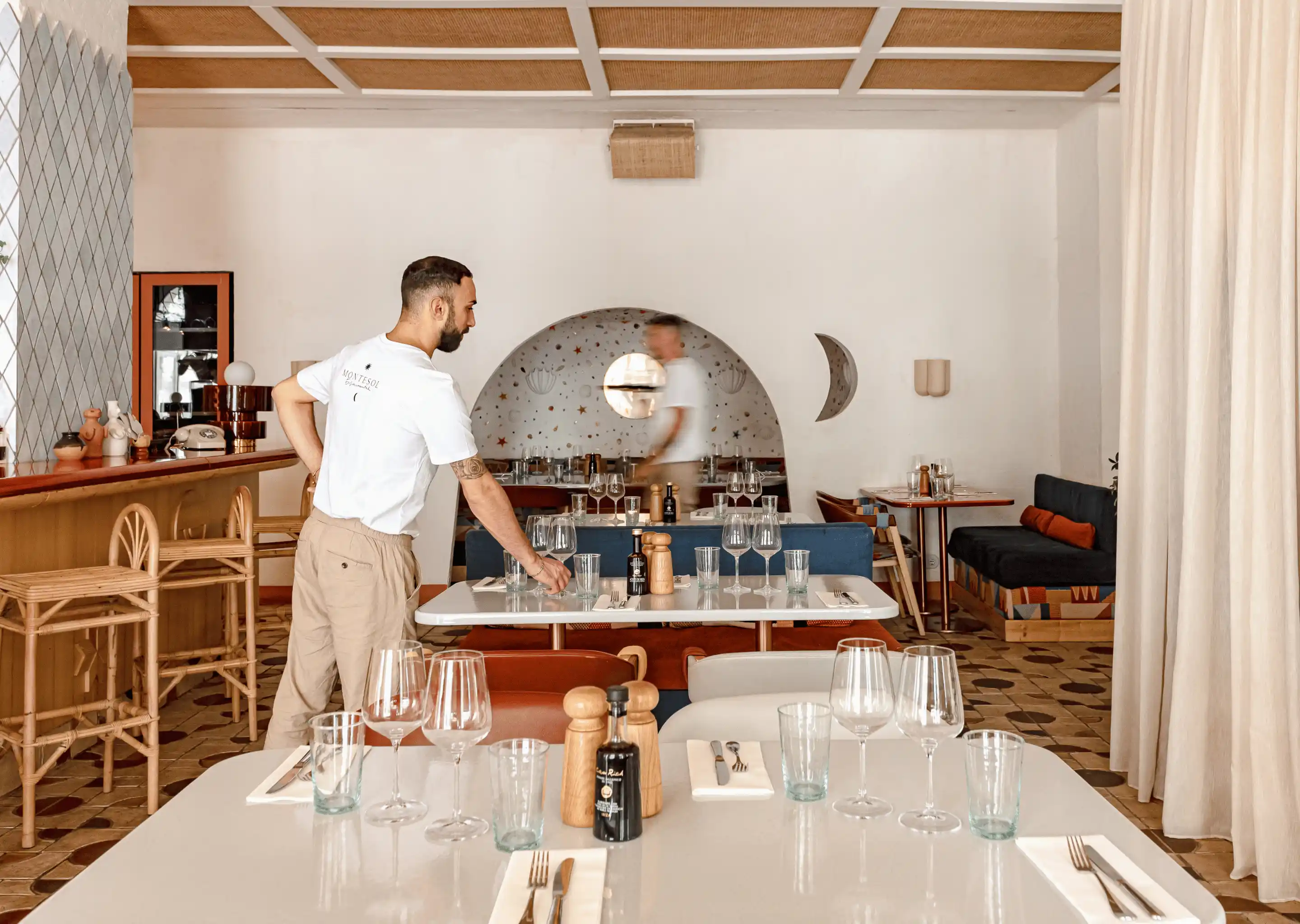 Man setting a table in a stylish restaurant with white walls, wooden ceiling, and patterned floor tiles.