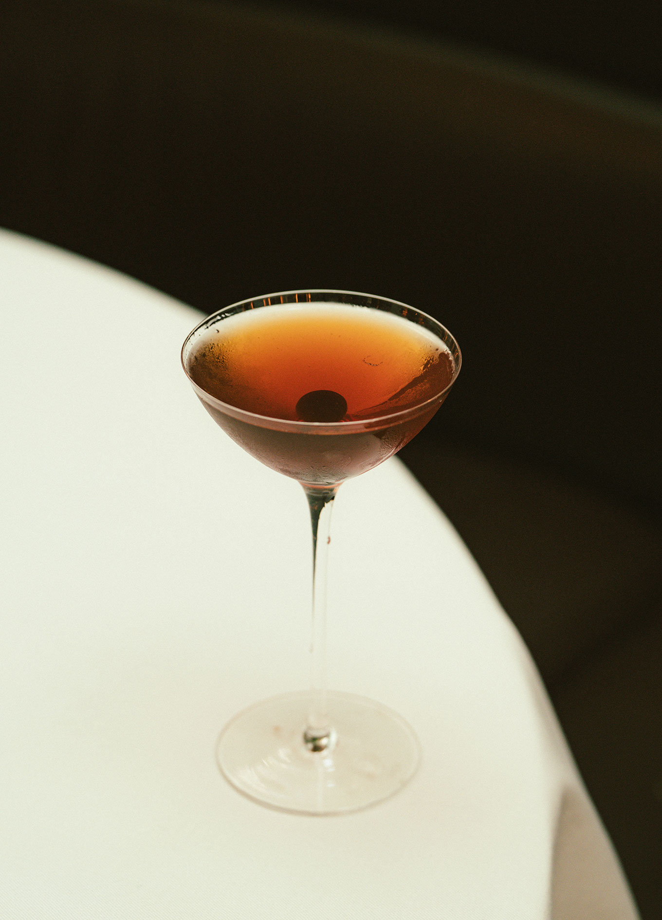 Cocktail in a stemmed glass with a reddish-brown liquid and a dark cherry garnish on a white tablecloth.