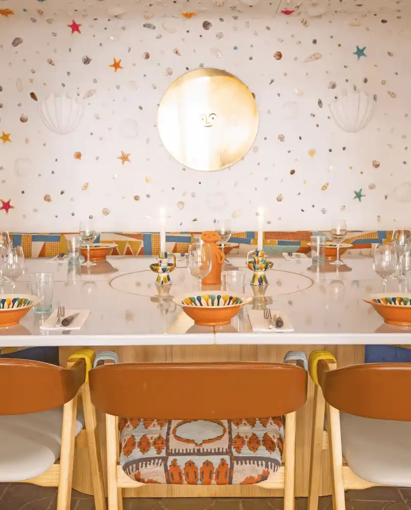 A dining table set with colorful bowls, glasses, and utensils in a room with a decorative wall featuring starfish, shells, and a round mirror with a face design.