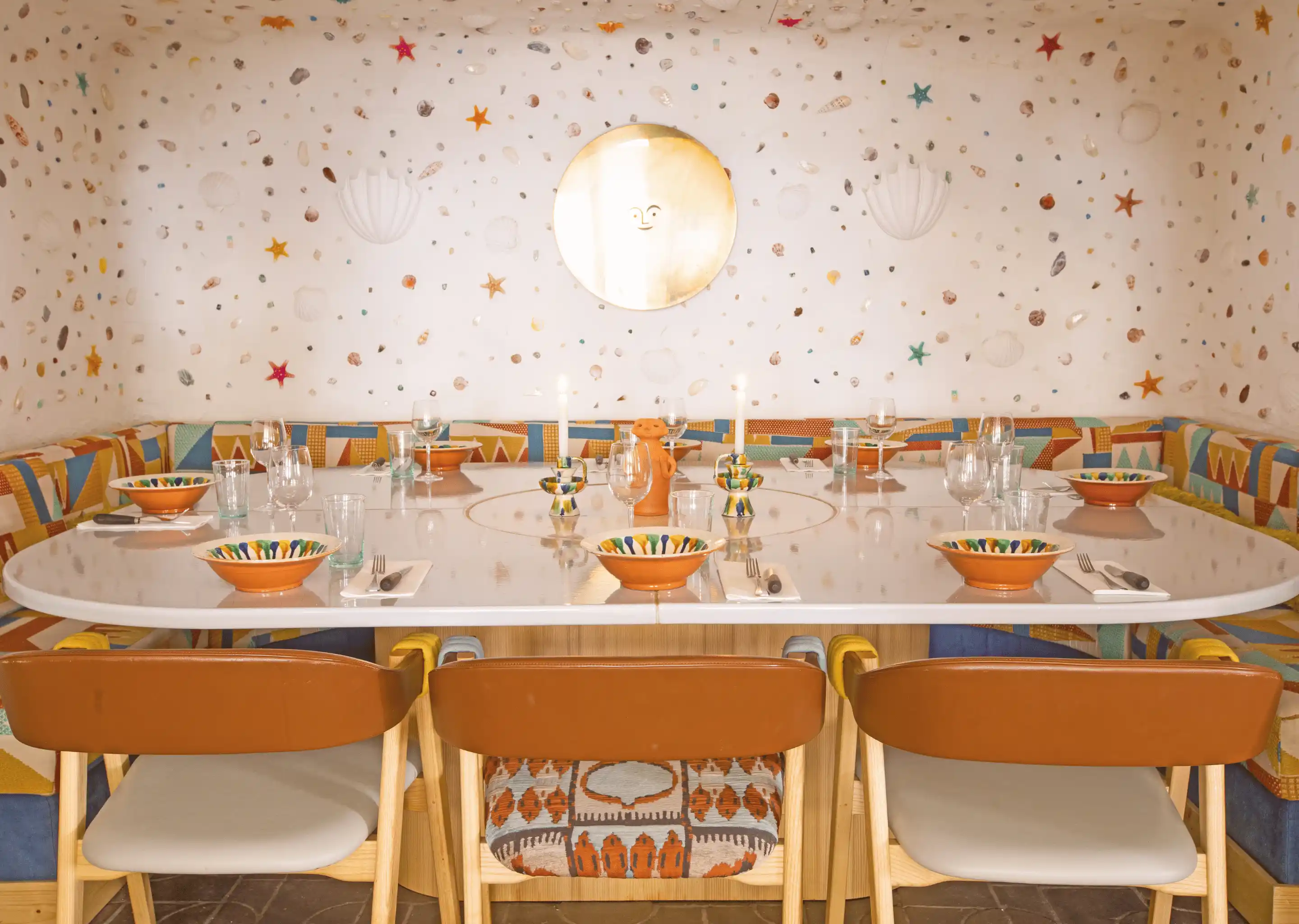 Table set for six in a room with colorful patterned bench seating and a wall decorated with seashells and starfish motifs, centered with a round golden mirror featuring a smiling face.