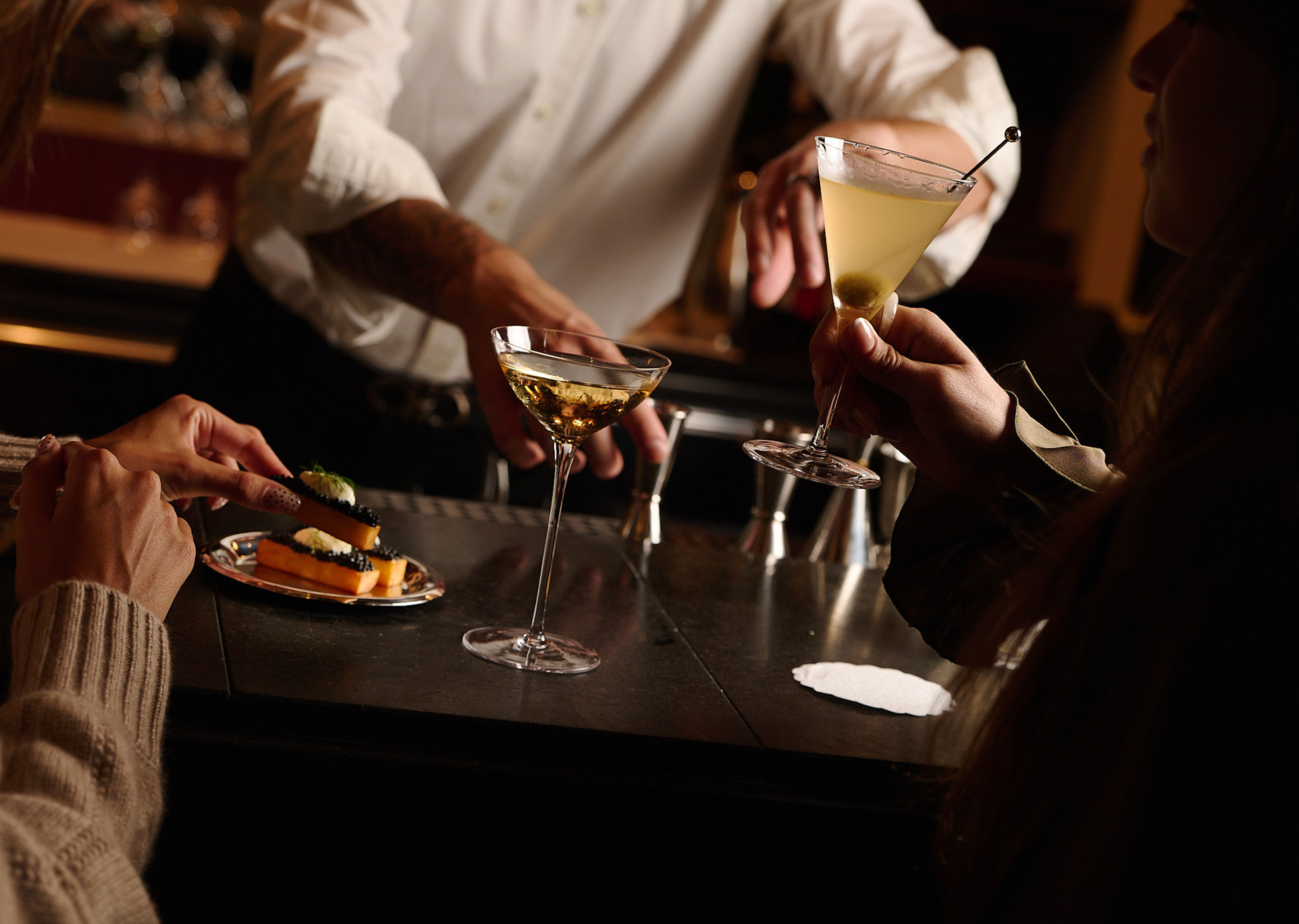 Bartender serving cocktails to two people at a dimly lit bar with an appetizer plate on the counter.