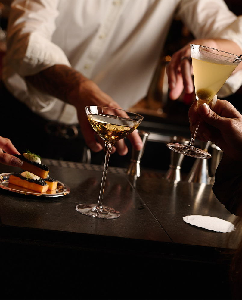 Two cocktail glasses on a bar counter, one held by a hand, with a bartender's tattooed arm reaching out and a plate of appetizers nearby.